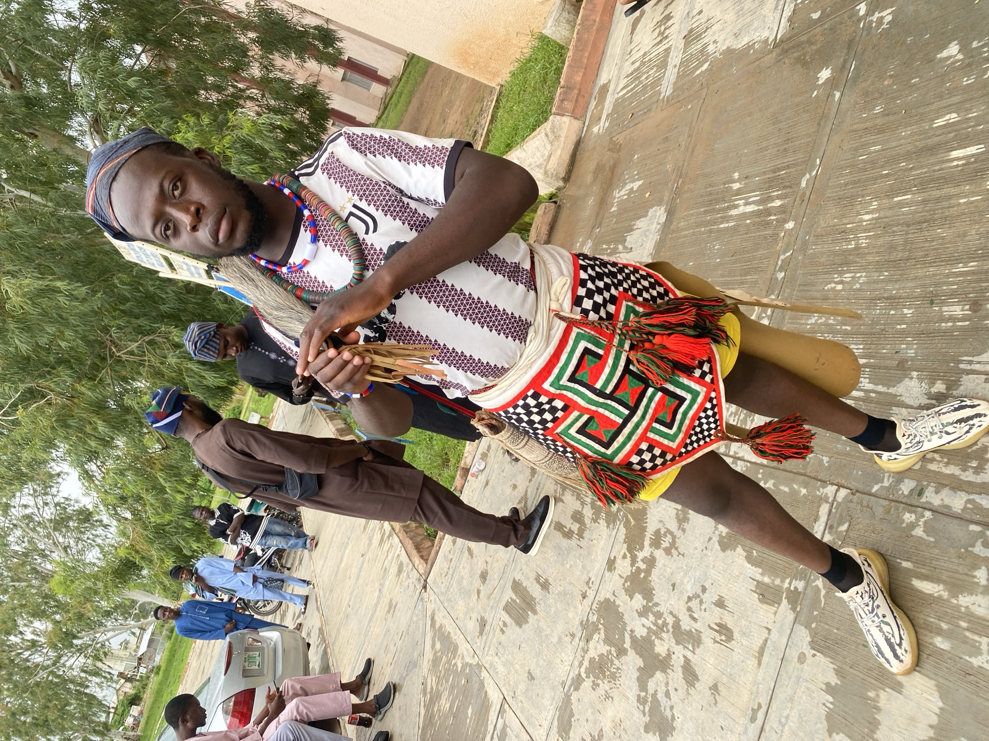 The image depicts a group of young men outdoors, appearing to be in a public space in Rafin Par, Nigeria. The foreground is dominated by a young man dressed in traditional attire, including a vibrantly patterned skirt, a striped shirt, and a beaded necklace. He is holding a bundle of what appear to be wooden implements. In the background, other men are visible, some in casual clothing and others in traditional garments, standing near parked vehicles and trees. The ground is paved with concrete, and there are indications of some dampness, possibly from recent rain. The overall atmosphere suggests a gathering or an event taking place.