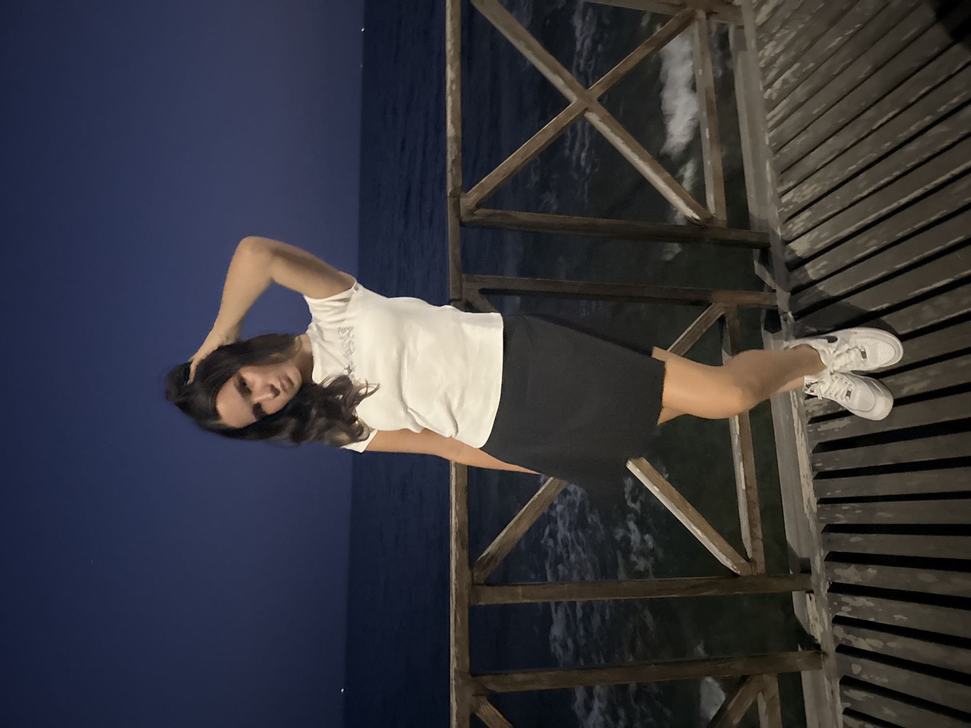 A young woman with long, dark hair stands on a wooden pier at night, with the sea and dark blue sky in the background. She is wearing a white t-shirt and a black skirt, with white sneakers. Her right hand is raised to her head. The pier has a wooden railing. The location is Agios Tychona Community, Cyprus.