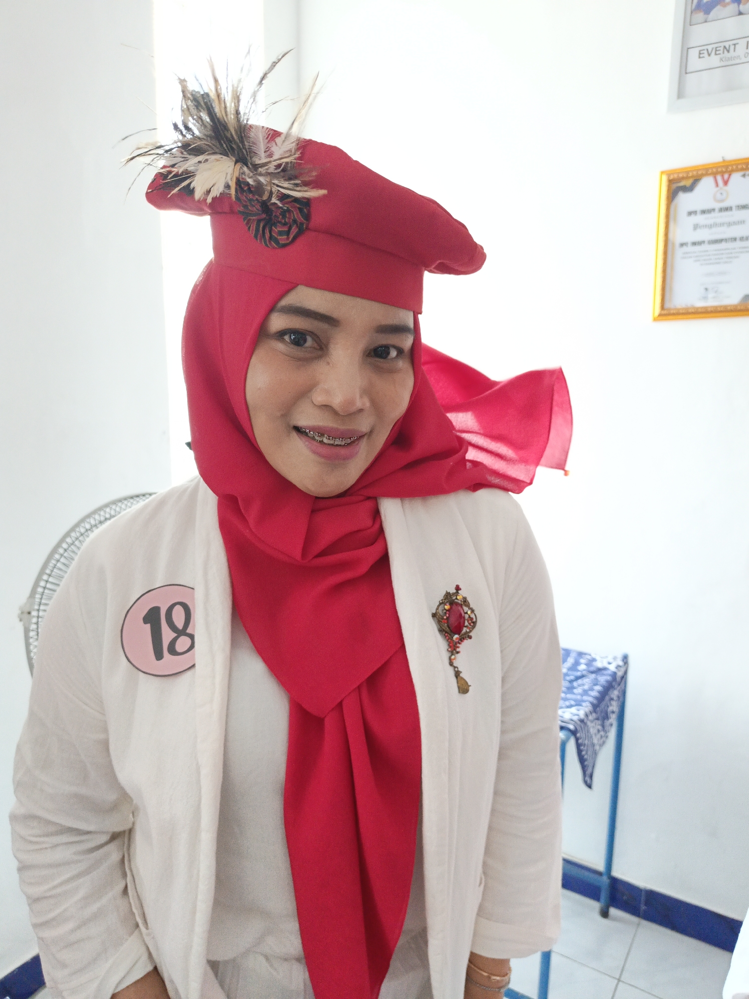 A medium shot shows a woman wearing a red hijab and a white jacket. She has a red hat with feathers on her head and a red brooch on her jacket. A pink circle with the number "18" is pinned to her jacket. The background is a white wall with framed certificates and a blue and white patterned table. A fan is visible on the left side of the image. The image appears to be taken indoors in Klaten, Indonesia, as suggested by the framed certificate mentioning "Klaten, 09."