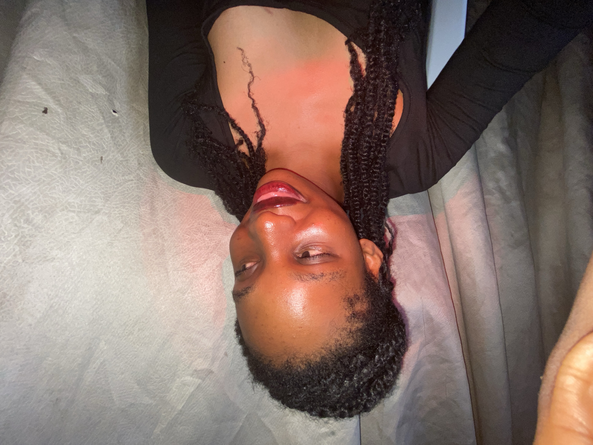 A person is lying on a surface, possibly a bed or couch, indoors. The individual has dark hair in braids and appears to be wearing a dark-colored, long-sleeved top. The lighting is somewhat dim with a possible red or pink hue casting on the skin. The surface they are lying on has a textured appearance. There is a curtain in the background, and a hand is present in the image. The location is Unknown, Unknown.