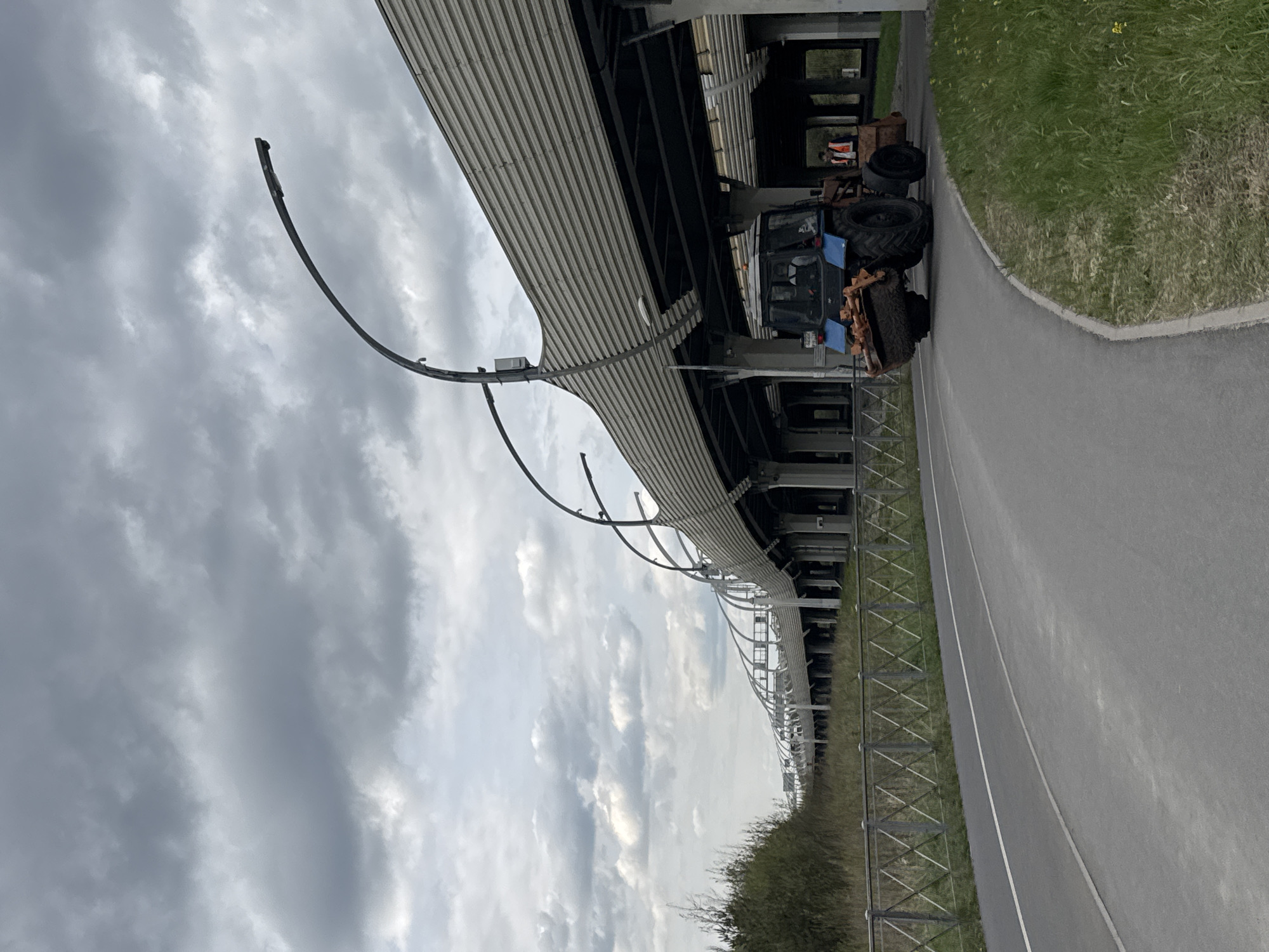 The image shows a cloudy sky above a section of the Igora Drive Formula 1 race track near Saint Petersburg, Russia. The track's curved, covered structure dominates the upper portion of the photo, with metal supports and a corrugated roof visible. Below the track structure, a service vehicle, possibly a tractor, is parked on the asphalt surface. A metal fence runs along the edge of the track, separating it from a grassy area with some bushes. The photo is taken from a low angle, emphasizing the height and scale of the race track infrastructure.