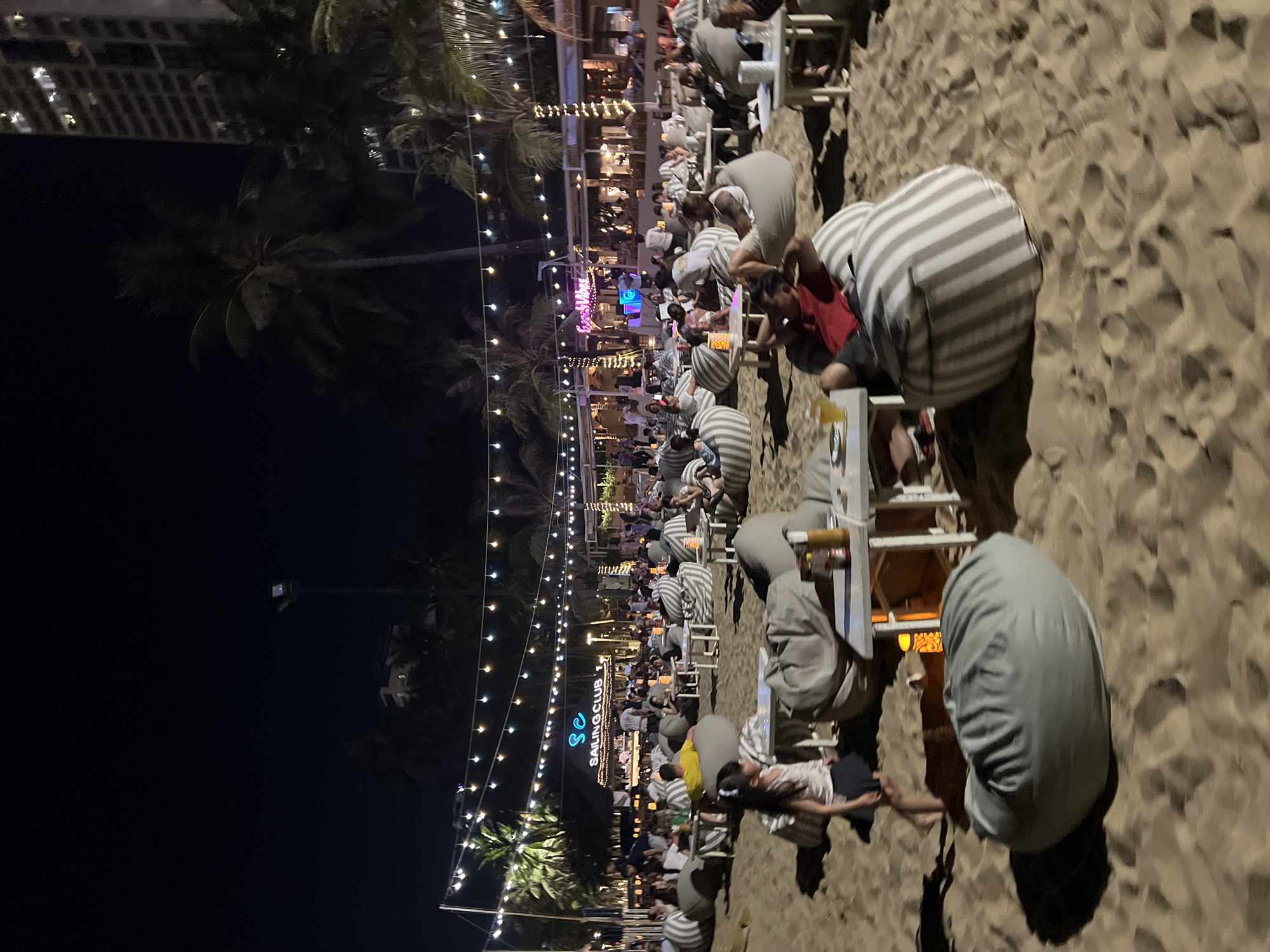 The image shows a nighttime scene at Sailing Club in Nha Trang Ward, Vietnam. Numerous people are seated at tables on a sandy beach, illuminated by string lights and the club's signage. Most patrons are sitting in chairs with gray and white striped cushions. Some are wearing casual clothes, and a few have beverages on their tables. The surrounding environment includes palm trees and a multi-story building in the background.
