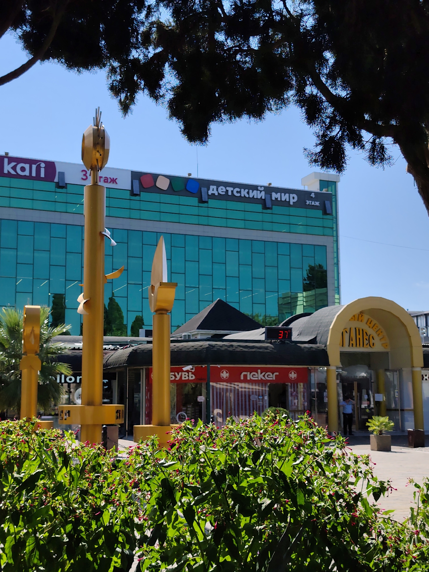 This image shows the "Detsky Mir" (Children's World) shopping center in Sochi, Russia, on a sunny day. A modern building with a distinctive turquoise glass facade is visible, adorned with signs in Russian, including "Detsky Mir" and "4 Etazh" (4th floor). In the foreground, a cluster of lush green bushes with small red flowers partially obscures the view of the shopping center's lower levels. Two prominent golden sculptures, resembling stylized plants or arrows, stand in front of the building. One sculpture is taller and more elaborate, featuring several curved elements and a pointed top. Another, shorter sculpture is visible to its left.
Beneath the "Detsky Mir" building, a covered walkway or entrance area is visible, housing various shops. Signs for "Rieker" and "Shov", as well as what appears to be a shoe store ("Obuv"), are identifiable. A digital display on the roof of this lower section shows the temperature as "37°". An arched entrance to a shopping center, labeled "Ganes" in yellow Cyrillic letters, is situated to the right of the "Detsky Mir" building. A person in a blue shirt and dark pants is standing near the entrance of "Ganes". Palm trees can be seen to the left of the golden sculptures. The sky is clear blue, and shadows suggest the sun is high in the sky.