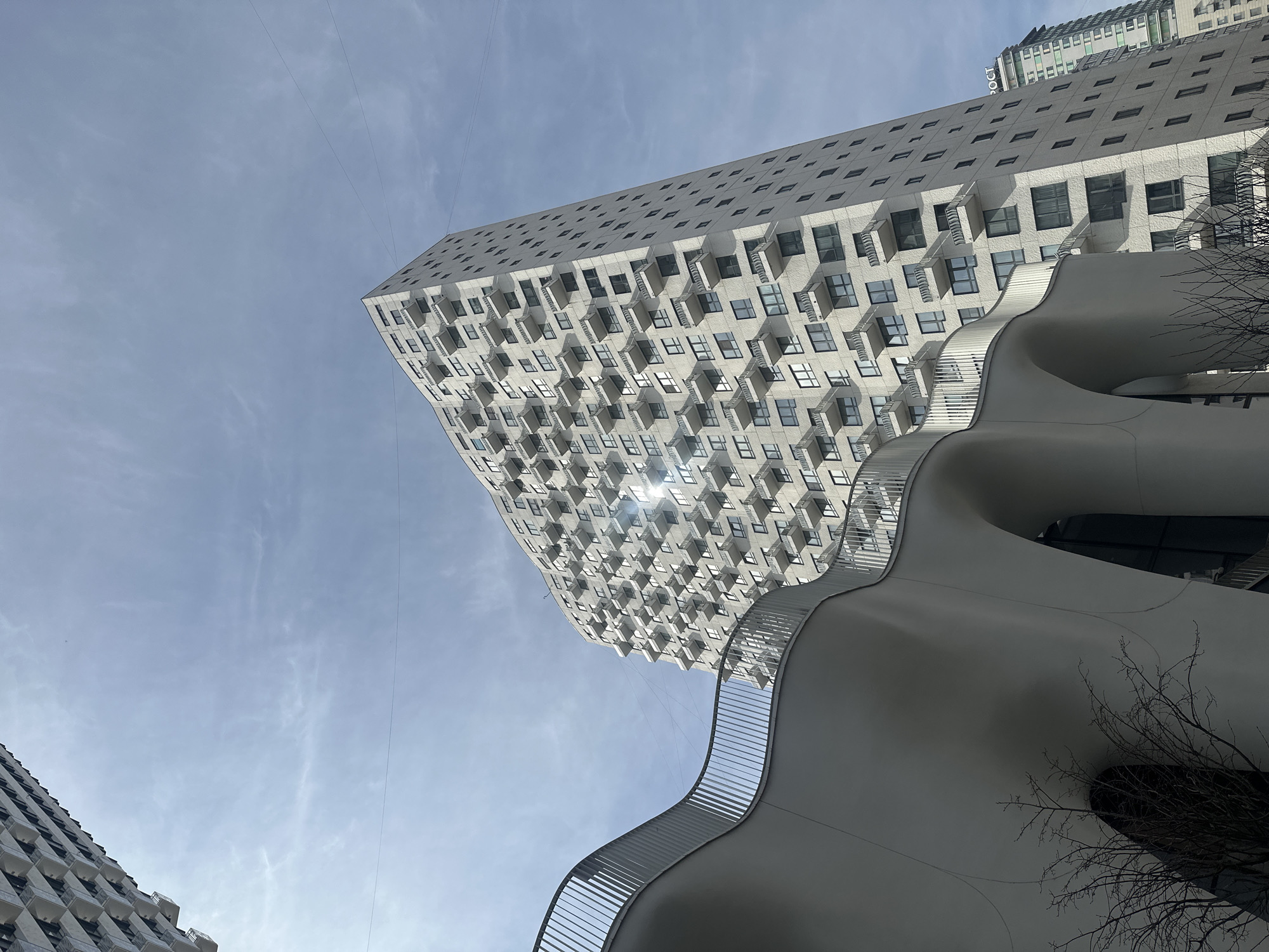 The image shows a low-angle view of two modern buildings in Moscow, Russia. The taller building in the background features a geometric design with numerous protruding balconies. Sunlight reflects off some of the windows. In the foreground, a smaller, white building with smooth, curving walls and a metallic railing is visible. The sky is pale blue with thin wisps of clouds. There are bare branches from a tree visible to the right. Power lines are visible against the sky.