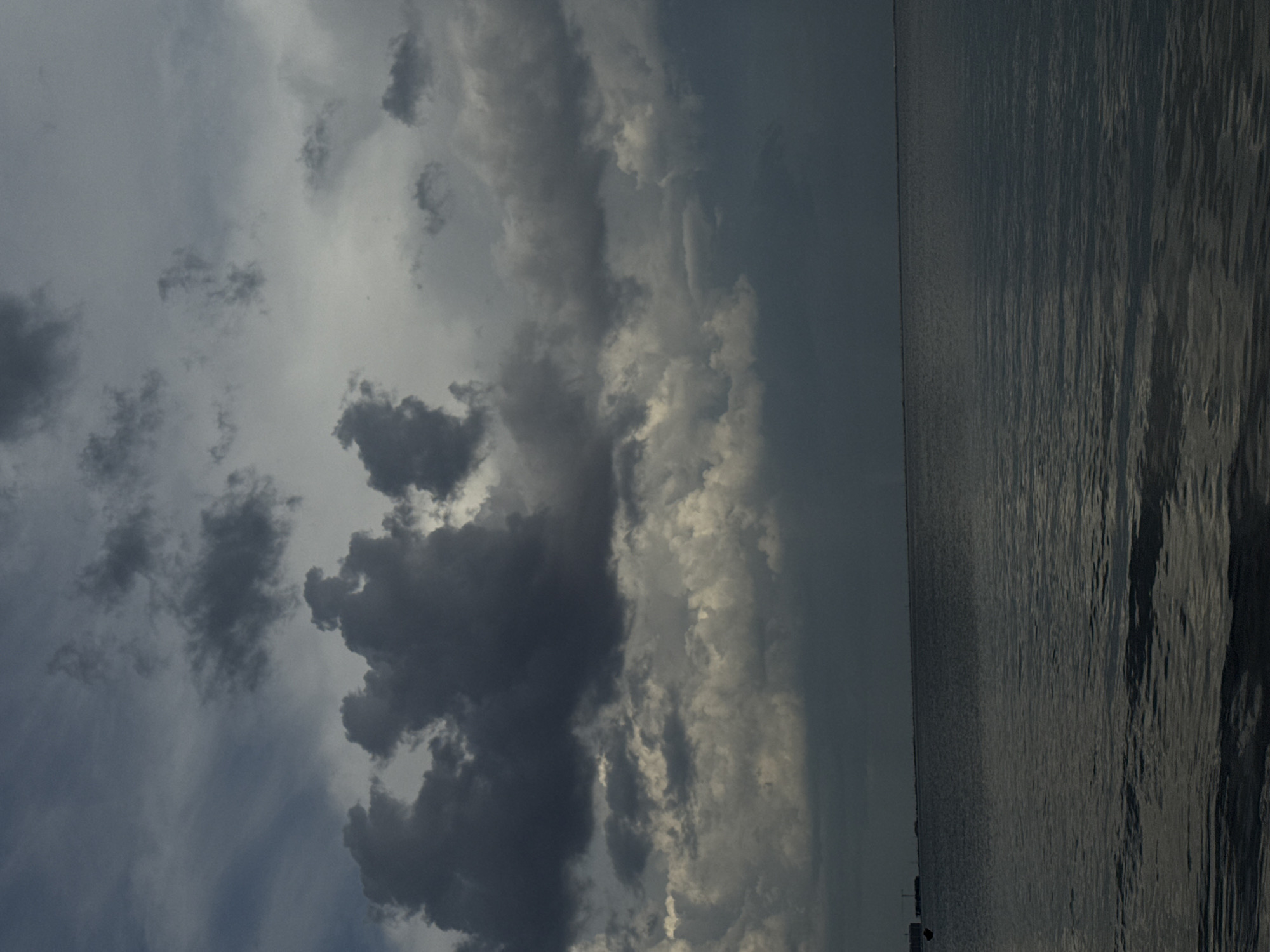 The image shows a body of water meeting a cloudy sky. The water is grey and rippled, suggesting a slight breeze. The sky is filled with dark, heavy clouds, indicating possible inclement weather. The scene evokes a sense of calmness or melancholy. The location is the city of Yevpatoriya, Ukraine.