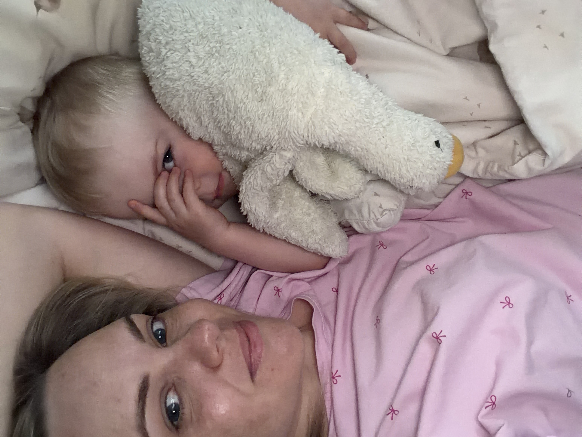 A fair-skinned woman and a young child with blond hair lie on a bed covered with pink bedding. The bedding has a bow pattern embroidered on it. The child is hugging a white plush toy in the shape of a goose. The child is looking directly at the camera with his right hand near his face. The woman is also looking at the camera. The scene appears to be indoors, presumably in a bedroom. The location is unknown, there is no information indicating that this photo was taken in the city of Votkinsk, Russia.