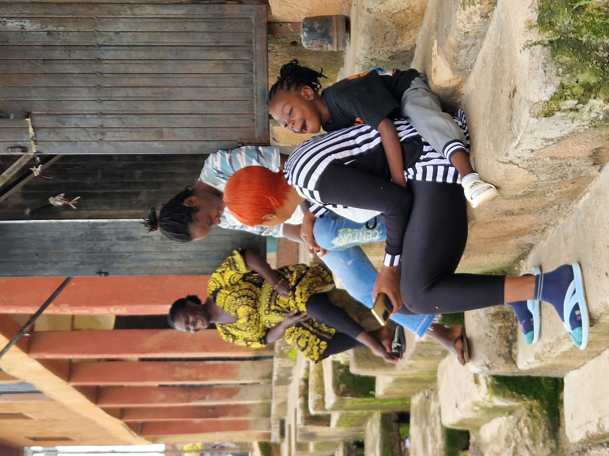 In an outdoor setting that appears to be the entrance to a building or home, likely in a village or rural area, a group of people are gathered on rough-hewn concrete steps. The steps are weathered and partially covered in green moss, suggesting an older or possibly neglected structure. Behind the steps, a weathered wooden door with vertical planks is visible, flanked by a rough concrete wall. To the left of the steps, an orange-tinted building with vertical support columns and a balcony railing is partially visible.
The central figures are two women and a child. One woman, wearing a vibrant yellow and black patterned dress and black leggings, sits on the steps, smiling broadly. Her feet are bare except for black flip-flops. Next to her, a woman with bright orange hair, dressed in a black and white striped top and black leggings, is seated, holding a mobile phone. She appears to be interacting with a child who is perched on her lap and leaning back, their face turned upwards in an expression of joy or laughter, eyes closed, mouth open. The child is wearing a dark t-shirt and grey pants. Another woman, wearing a blue and white striped t-shirt, is partially visible behind and to the left of the woman with orange hair, her posture suggesting she is also seated on the steps. A fourth individual, their lower body visible on the steps, is wearing black leggings and blue socks with white and blue sandals. The overall impression is a moment of casual social interaction on a warm day.