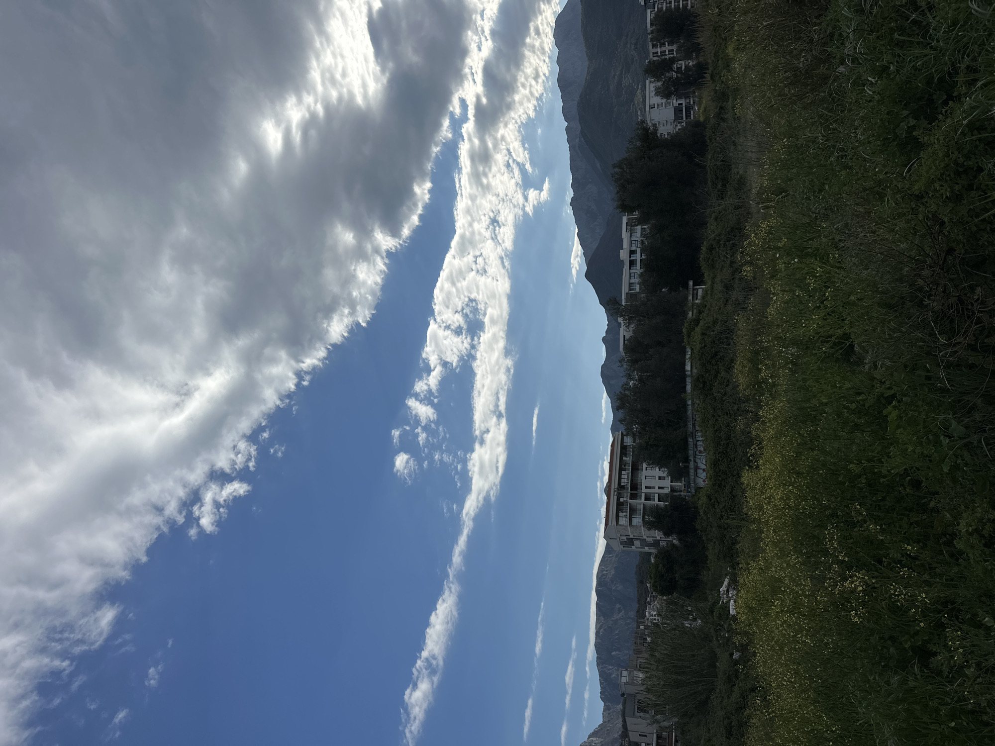 The image shows a landscape with a view of a mountain range and a partly cloudy sky in Šušanj, Montenegro. White buildings are visible on the lower slopes of the mountains. The foreground features vegetation.