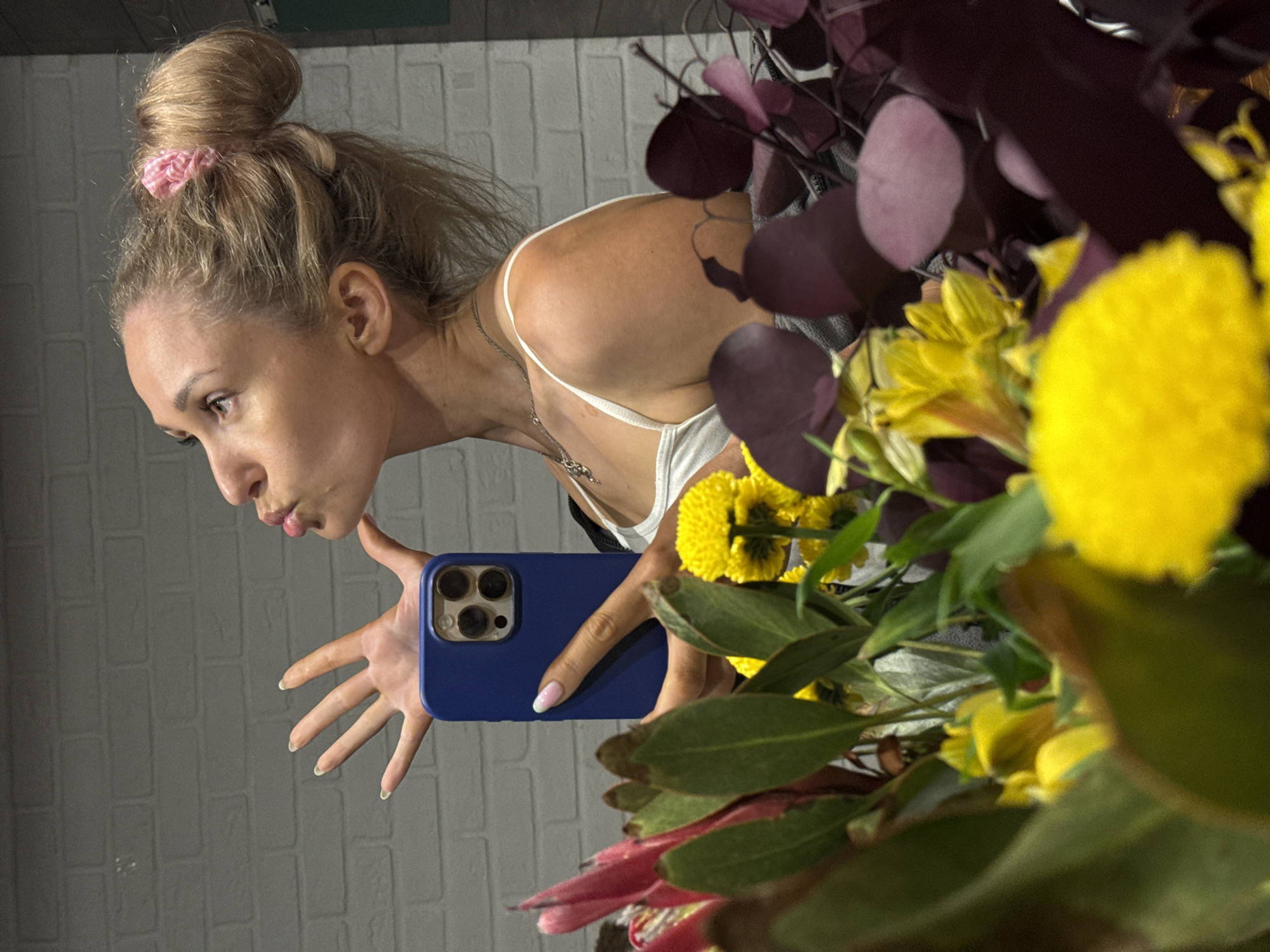 A woman is taking a mirror selfie, captured in profile, facing left. She has blonde hair styled in a high bun secured with a pink and white striped scrunchie. Her head is tilted slightly back, and her lips are pursed in a kissing gesture, with her eyes looking upwards. She wears a white spaghetti-strap top and a thin silver-colored necklace with two small pendants. Her left hand is extended forward with fingers splayed, revealing light-colored nails with pale tips. Her right hand holds a dark blue smartphone case, which covers a device with a triple-lens camera array and flash, consistent with an iPhone Pro model. The visible fingernail on her right hand features a pink base with a mint green tip. The immediate foreground is dominated by a dense floral arrangement, including various yellow flowers (some round, some trumpet-shaped), deep red/pink protea-like blooms, dark burgundy/purple circular leaves, and green foliage, partially obscuring the lower half of the woman. The background consists of a textured white painted brick wall. There are no discernible visual cues within the image to indicate a specific location in the city of Chaikovsky, Russia.