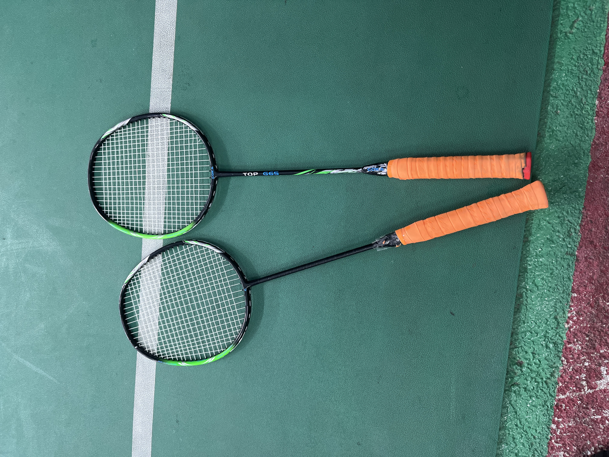 Two badminton rackets are lying on a green badminton court with an orange grip on the handle. The rackets are oriented approximately parallel to each other. One racket is partially resting on a white boundary line. The environment appears to be indoors, possibly a gymnasium. This is in a city in Bac Nha Trang Ward, Vietnam.