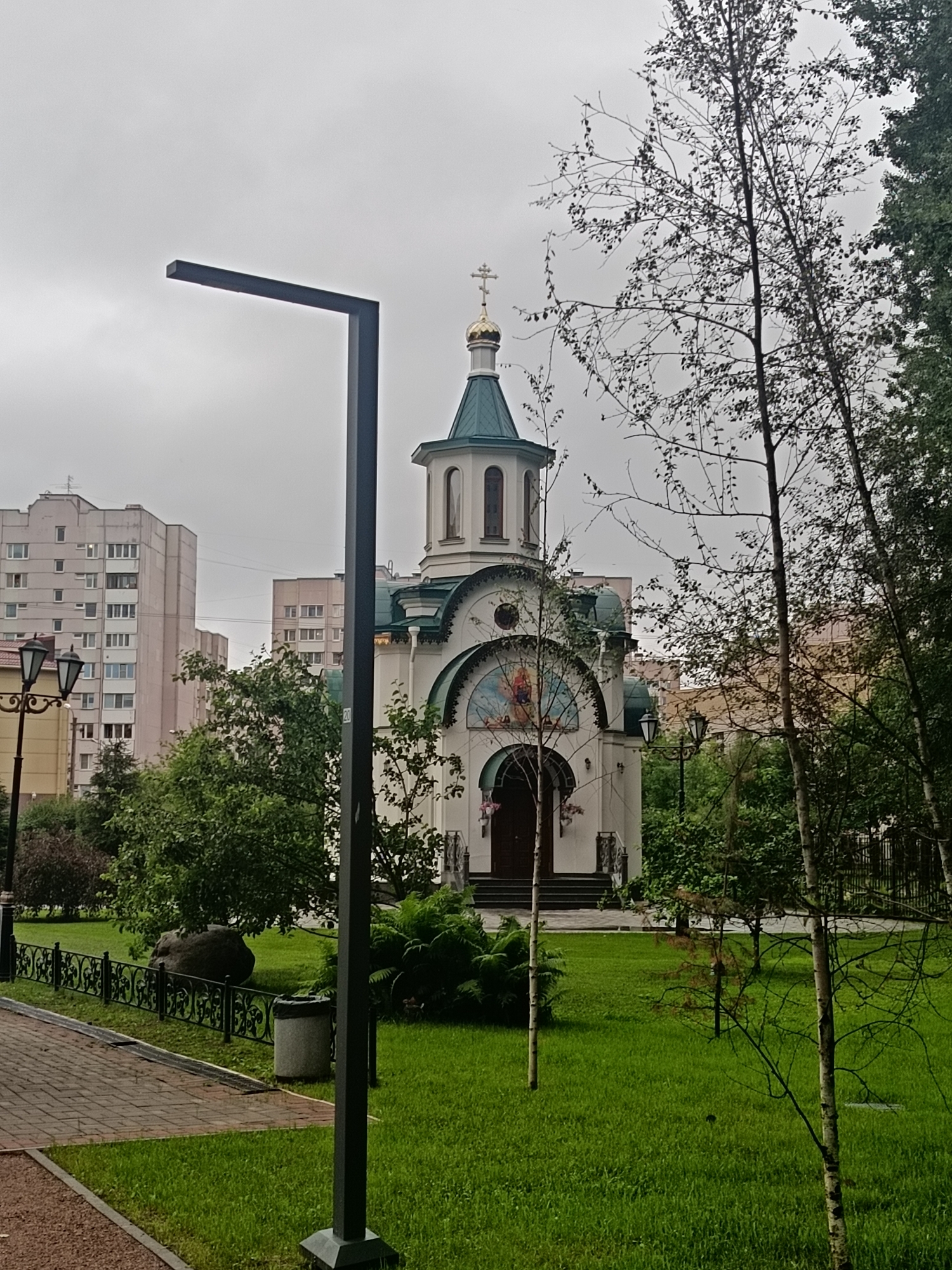 The image depicts the Church of the Holy Righteous Simeon the God-receiver and Anna the Prophetess, located in Saint Petersburg, Russia, in the area of Murino. The church is a white building with a green roof and a golden dome topped with a cross. It is situated in a park-like setting with a well-maintained lawn and various trees. A modern black lamppost stands in the foreground, to the left of the church. In the background, multi-story residential buildings are visible, suggesting an urban environment. The sky is overcast, indicating a cloudy day. There are no people visible in the image.