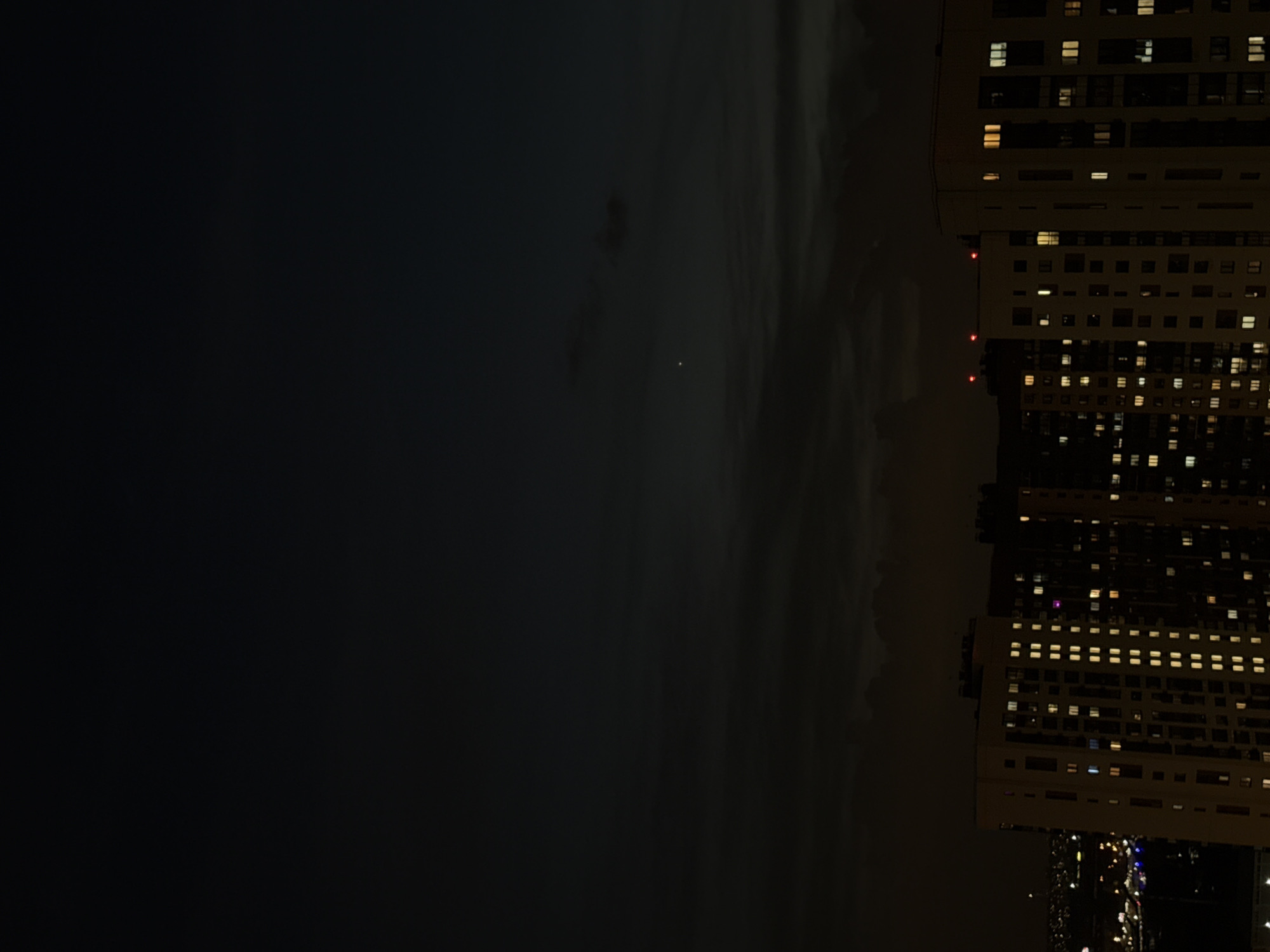 At night, a tall building is located in Lyubertsy, Russia. The building is illuminated, with many windows lit up. The sky is dark and cloudy.