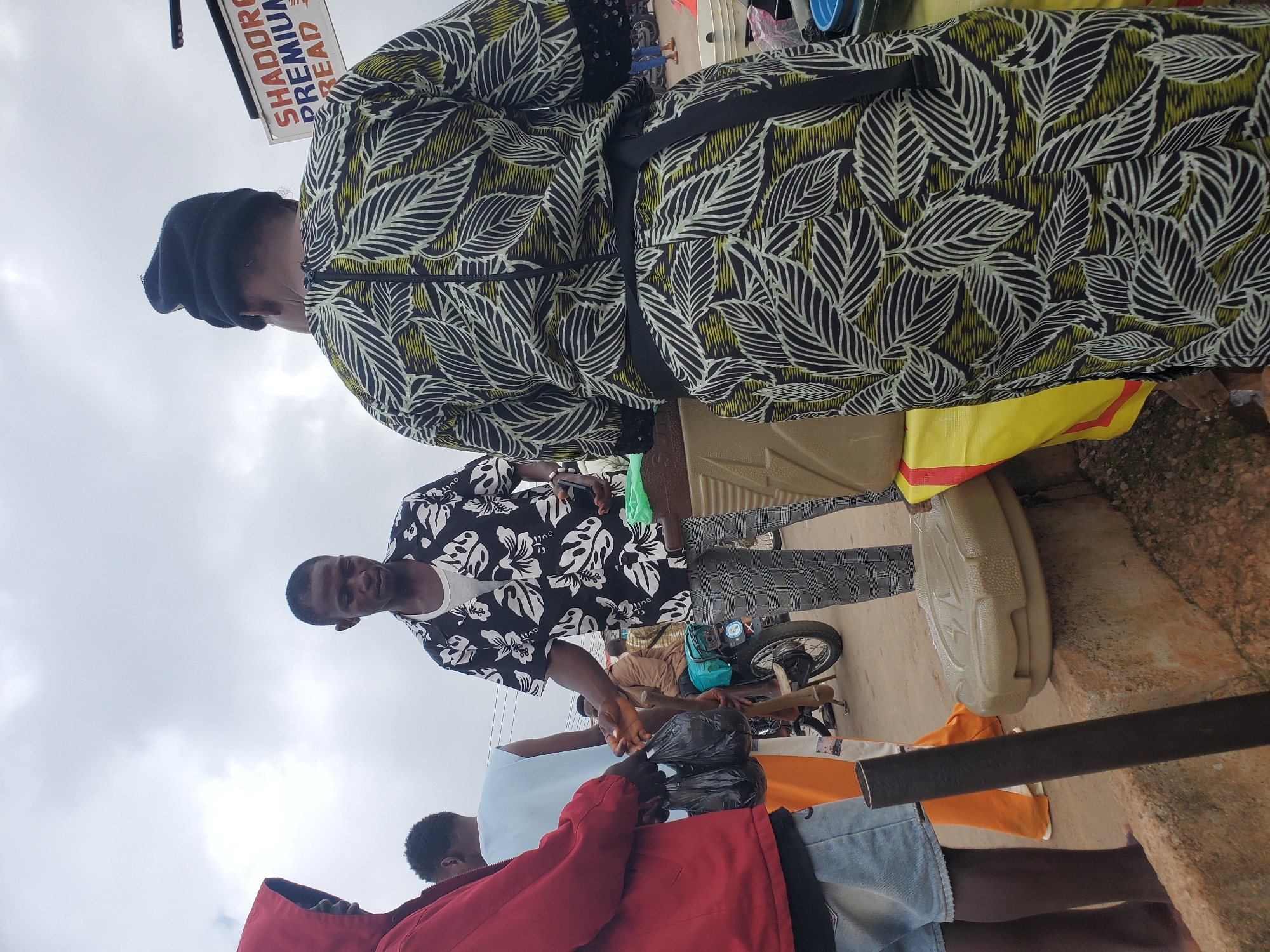 In Gwong, Nigeria, an outdoor scene unfolds under an overcast sky. Three men and one person wearing a black beanie are the central figures. The man on the far left wears a red hooded jacket and blue denim shorts. Next to him, a man in a light blue sleeveless shirt stands behind another man. This second man, wearing a black and white floral shirt and checkered trousers, is engaged in an exchange with the person in the beanie. This individual, whose back is to the camera, is dressed in a patterned green, black, and white dress and wears a black beanie. The person in the beanie is interacting with the man in the floral shirt, who is holding a phone. In the background, a motorcycle with a person wearing a brown jacket is visible. Several containers are present, including a beige one on a stone ledge and a large yellow tarp spread out. A sign with the text "SHADORA PREMIUM BREAD" is affixed to a pole. The ground appears to be a dirt road.