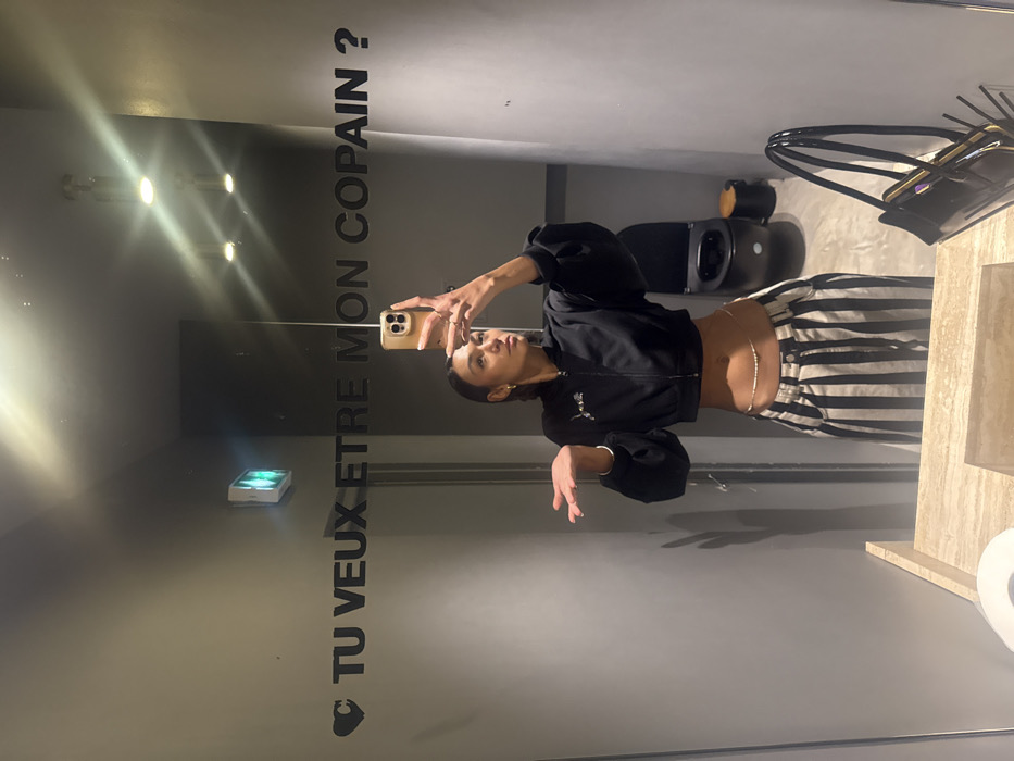 A woman is depicted taking a mirror selfie in a public restroom, identified as likely being at Pink Mamma restaurant in Paris, France, due to the prominent French text on the mirror. The subject has dark hair and wears a black cropped bomber jacket, black and white vertical striped wide-leg pants, and a beaded waist chain. She holds a light-colored smartphone in her right hand to capture the image, while her left hand is open with the palm facing upward. The mirror displays a black stylized heart followed by the French phrase "TU VEUX ETRE MON COPAIN ?" ("Do you want to be my boyfriend?") and reflects a green exit sign. The restroom features gray walls and a beige, veined stone countertop. A black handbag containing a smartphone rests on the countertop to the right. In the background reflection, a dark-colored toilet and a dark cylindrical waste bin are visible, illuminated by two ceiling-mounted light fixtures.