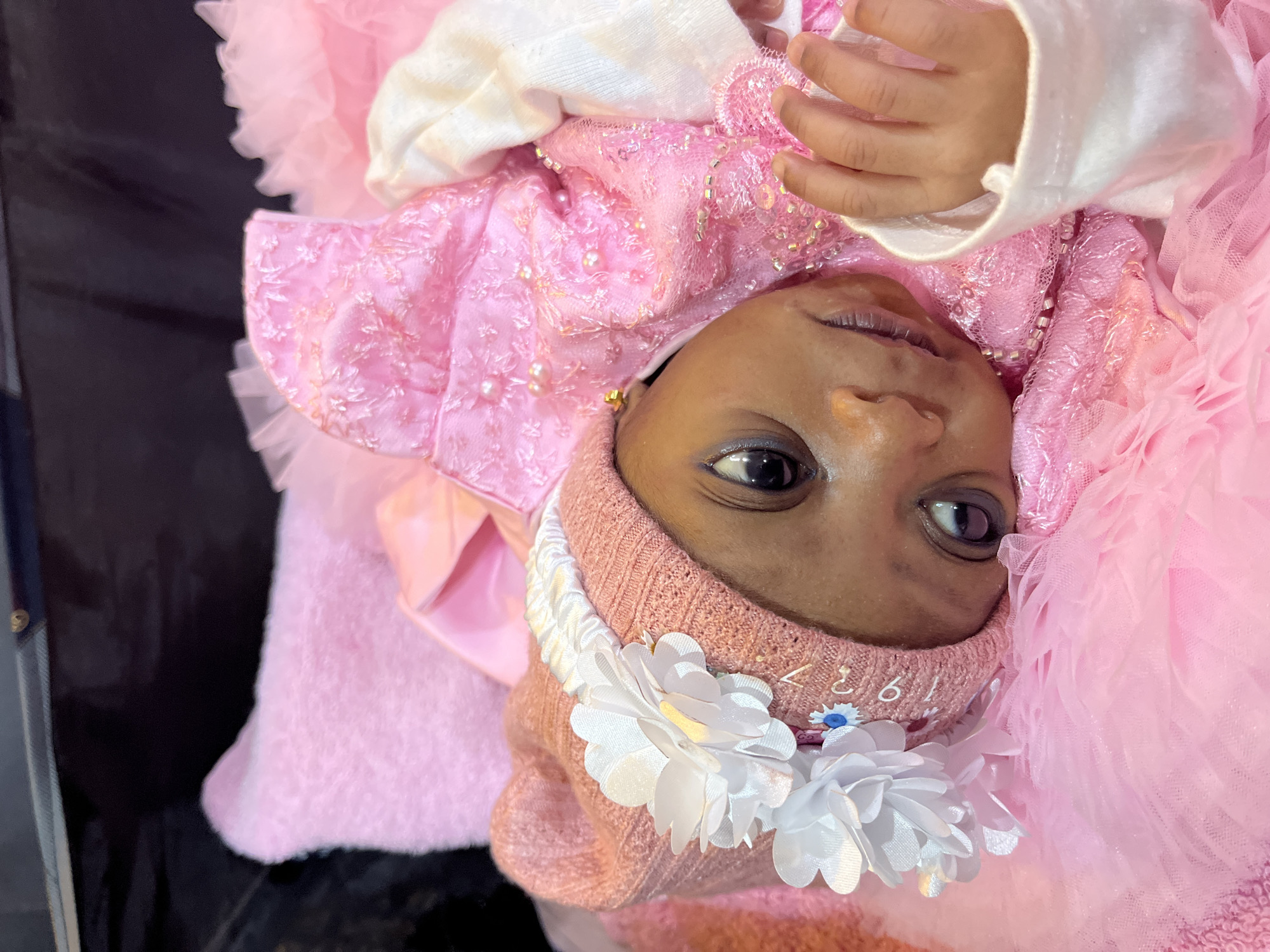 The image shows a close-up, high-angle view of a newborn baby girl lying down, adorned in a pink outfit. She is wearing a pink knitted hat with white floral accents and a white headband. Her dress is also pink, featuring intricate detailing with beads and sequins. The baby is positioned on a pink, fluffy surface, likely a blanket or crib, with layers of pink tulle fabric around her. Her eyes are open, and she appears to be looking towards the viewer. A small portion of her hand is visible on the right side of the frame, with her fingers slightly curled. The background is dark and out of focus, providing no discernible environmental clues. The overall impression is that of a carefully dressed infant, possibly for a special occasion. There is no specific information within the image to indicate the location in Jos, Nigeria.
