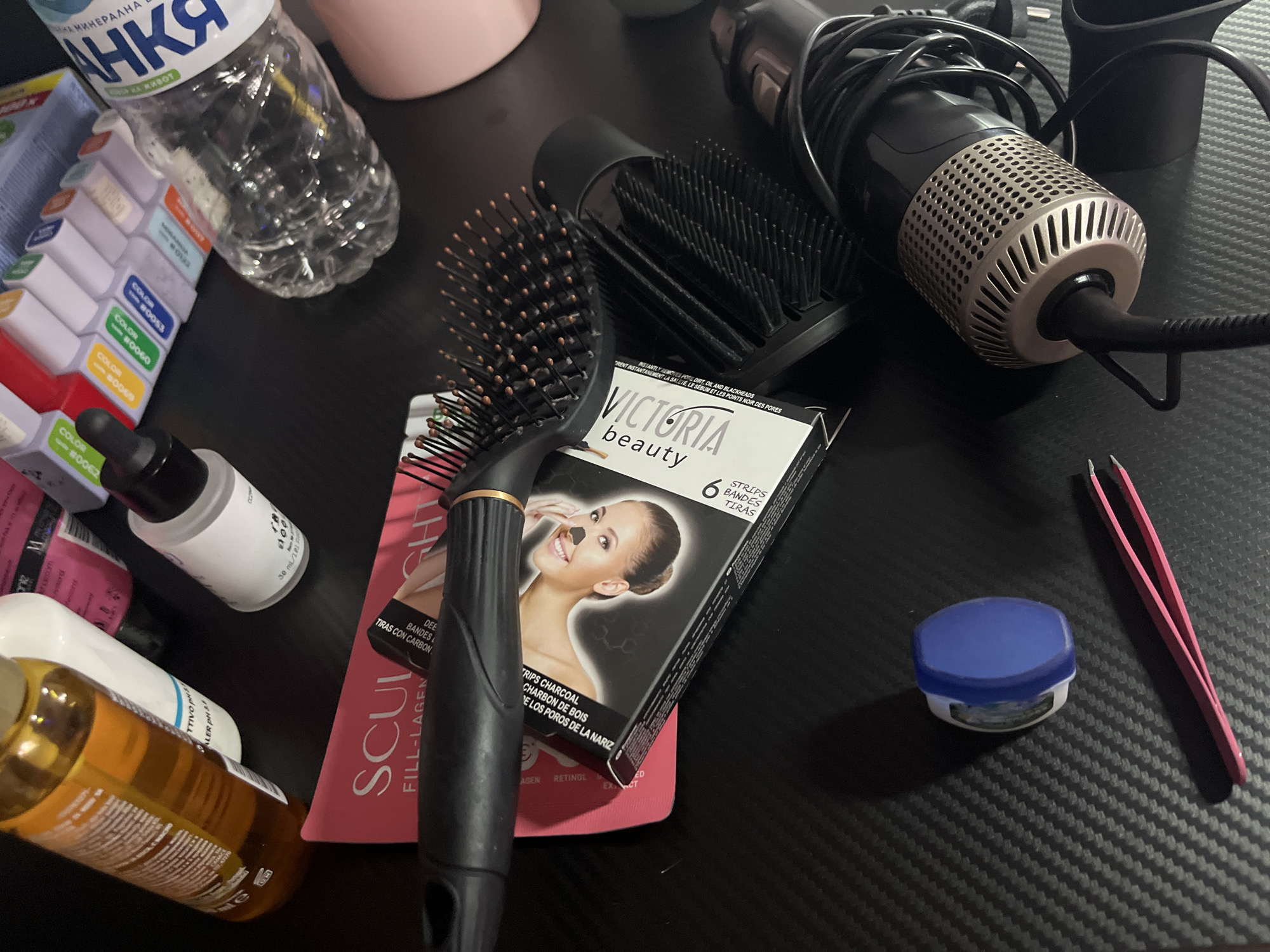 The image shows a collection of personal care and cosmetic items scattered on a dark, possibly carbon fiber textured surface. There is a water bottle with a Bulgarian brand name ("Анка минерална вода за живот"), suggesting the location may be in Bulgaria, and more specifically, Varna, considering this brand is available in that city. Items include:
* A water bottle with the brand name "АНКА".
* A set of nail polish bottles with various colors.
* Two bottles of liquid, one clear and one amber-colored.
* Two hairbrushes.
* A hairdryer with a black and silver body.
* A box of Victoria Beauty charcoal nose strips.
* A pink facial mask packet.
* A small jar of Vaseline.
* A pair of pink tweezers.
The items are arranged haphazardly, suggesting they are in use or recently used. The dark surface and the array of items suggest this could be a personal grooming area in a home setting.