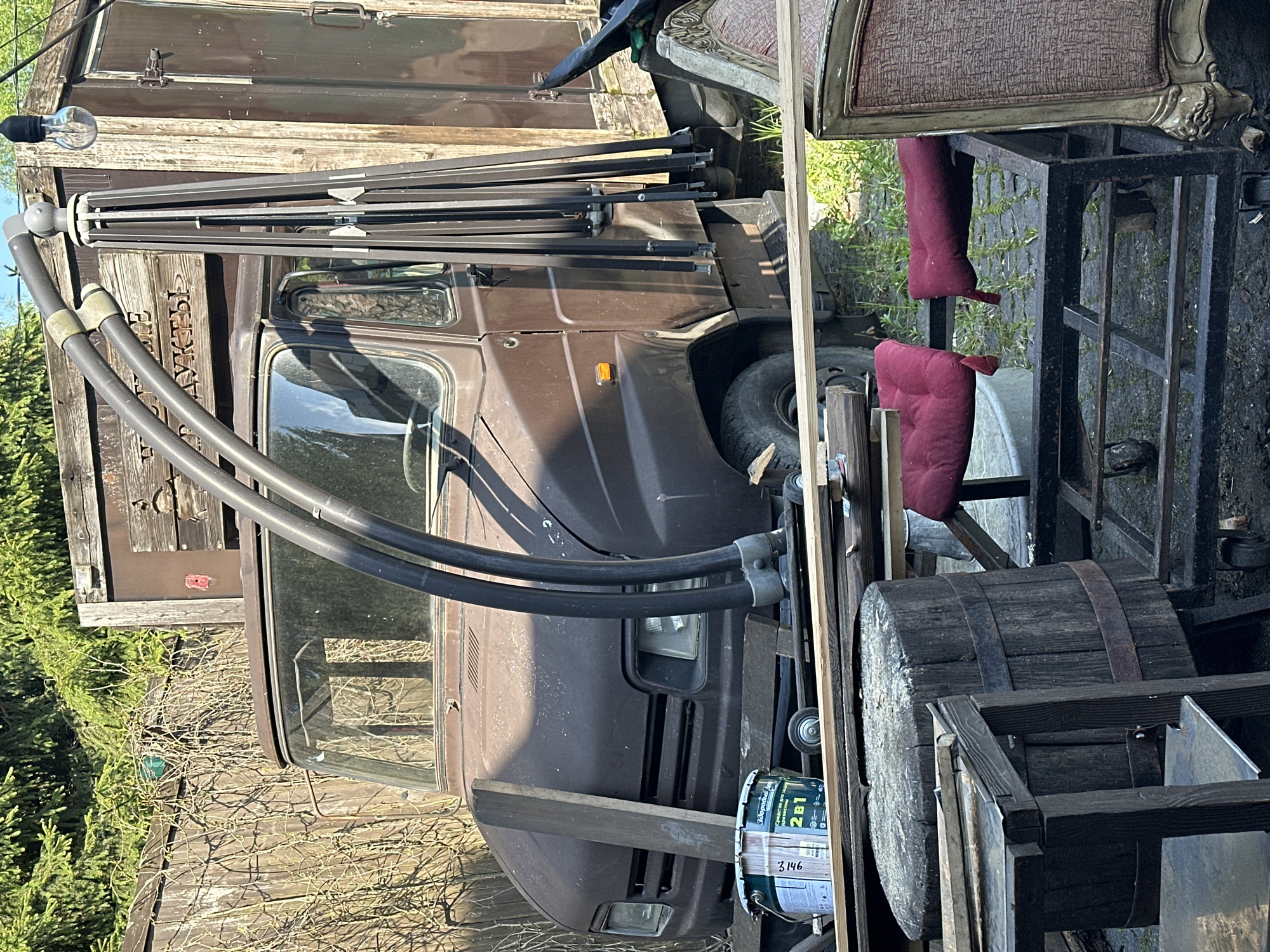 The image shows a collection of assorted objects outdoors. Visible are parts of a brown vehicle, a wooden barrel, furniture including a chair with red cushions, painting supplies, and wooden planks. A collapsible clothesline with an exposed lightbulb at the end is attached to a weathered wooden structure. The location is possibly Novo-Glagolevo, Russia, based on the Cyrillic text on the vehicle. The overall impression suggests a storage or work area.