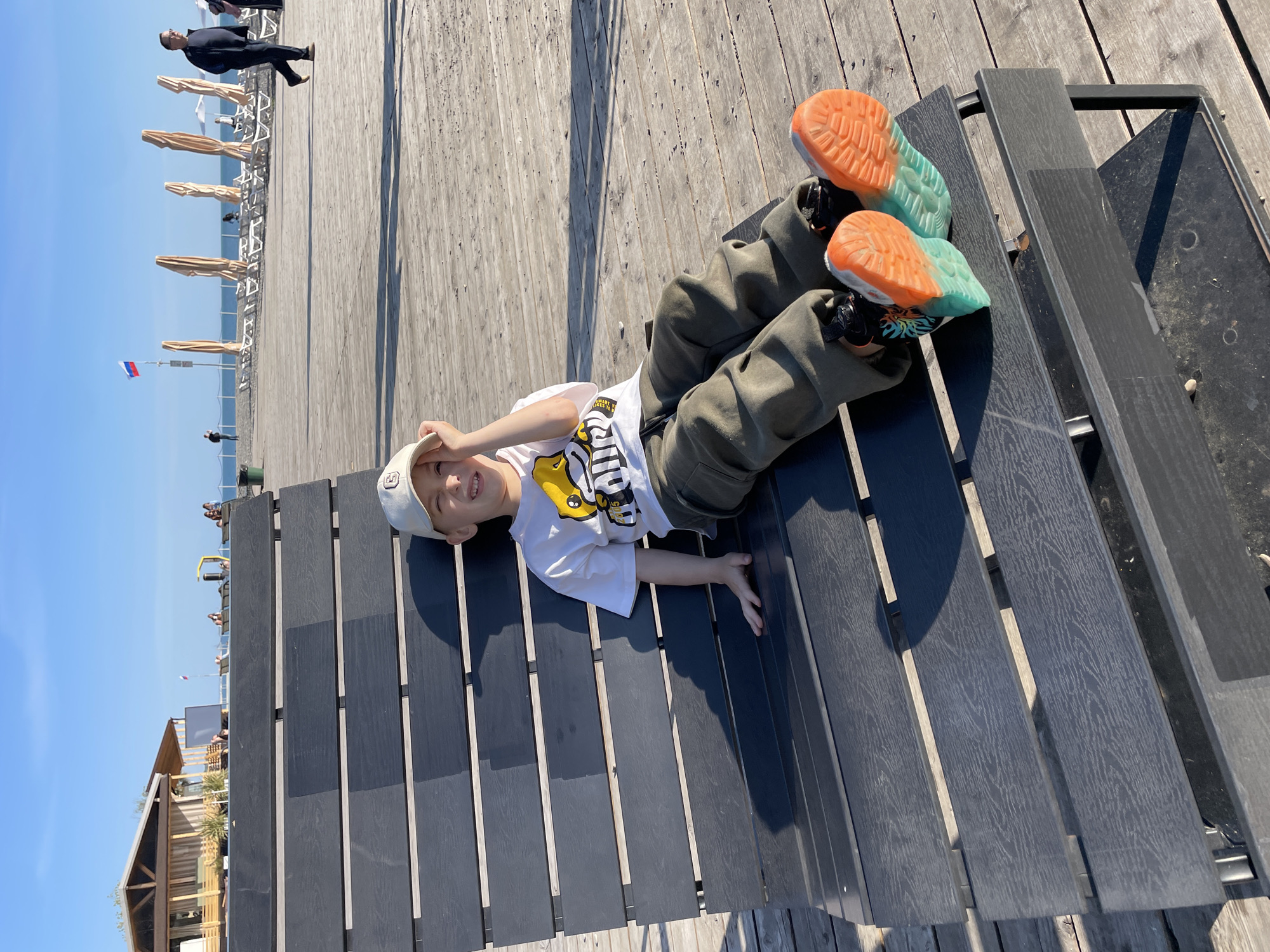 A boy in a white cap, a white t-shirt with a cartoon duck, and green pants is lying on a black bench. He is shielding his eyes with his hand and smiling. His shoes have orange and teal soles. The bench is made of black slats and has metal supports. The bench is on a wooden boardwalk. In the background, there is a beach with umbrellas, the sea, a Russian flag, and a building. A man in a black suit is walking in the background. The image was taken in Sochi, Russia.
