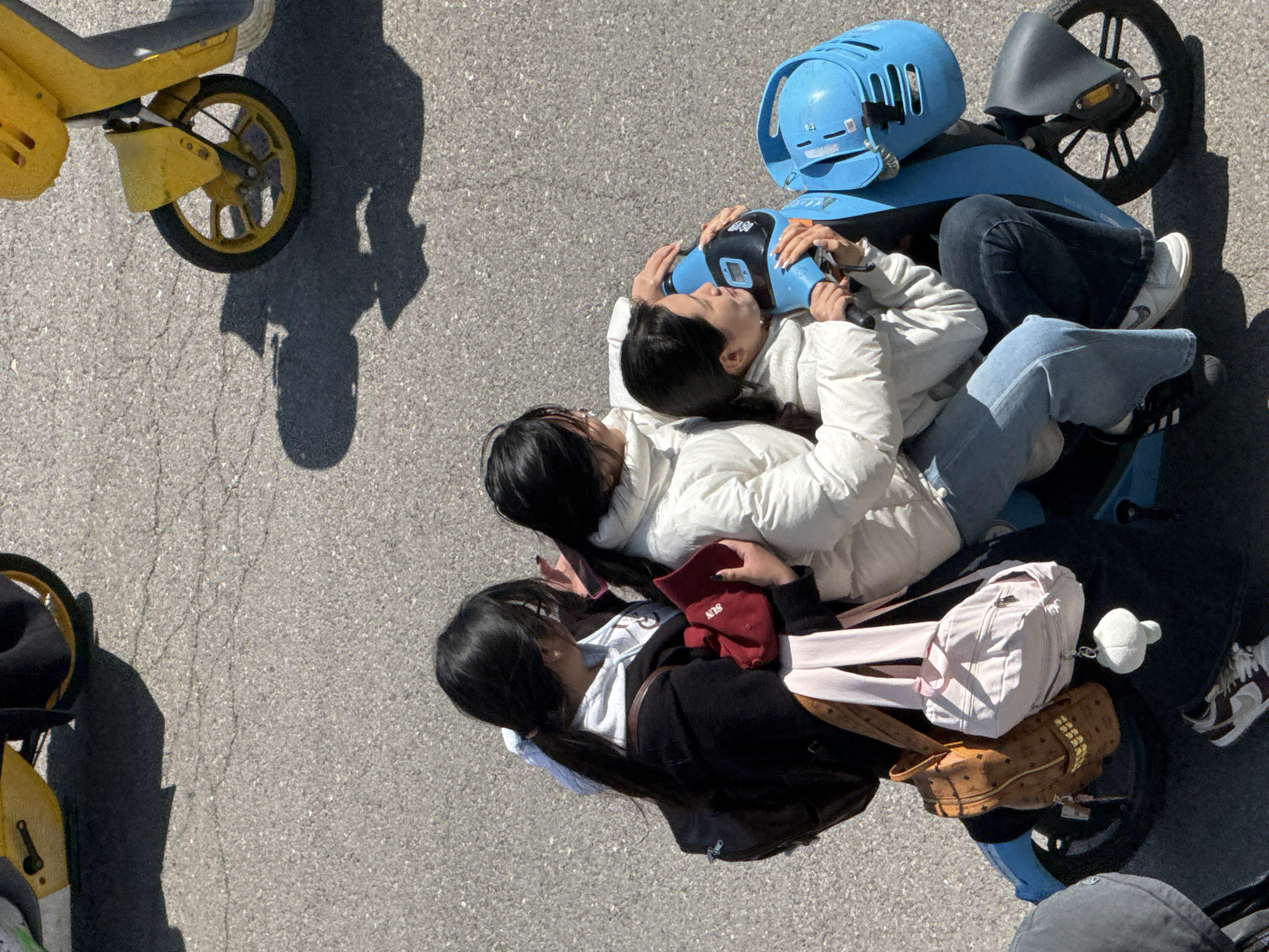 The image, taken from an elevated perspective, depicts three individuals interacting with two blue electric scooters and a yellow electric scooter on a gray asphalt surface, possibly in Zhongtun, China.
One individual is lying on top of another on one of the blue scooters, holding a blue helmet. The person below is wearing a white jacket and light-colored jeans with black and white sneakers. The person above is wearing a white jacket and dark denim jeans.
A third individual, wearing a dark jacket over a white top, is also on one of the blue scooters, amid several bags, including a brown bag, a white backpack, and a red bag. They are holding a smartphone.
A blue helmet sits atop one of the blue scooters. A yellow scooter is partially visible on the left side of the image.