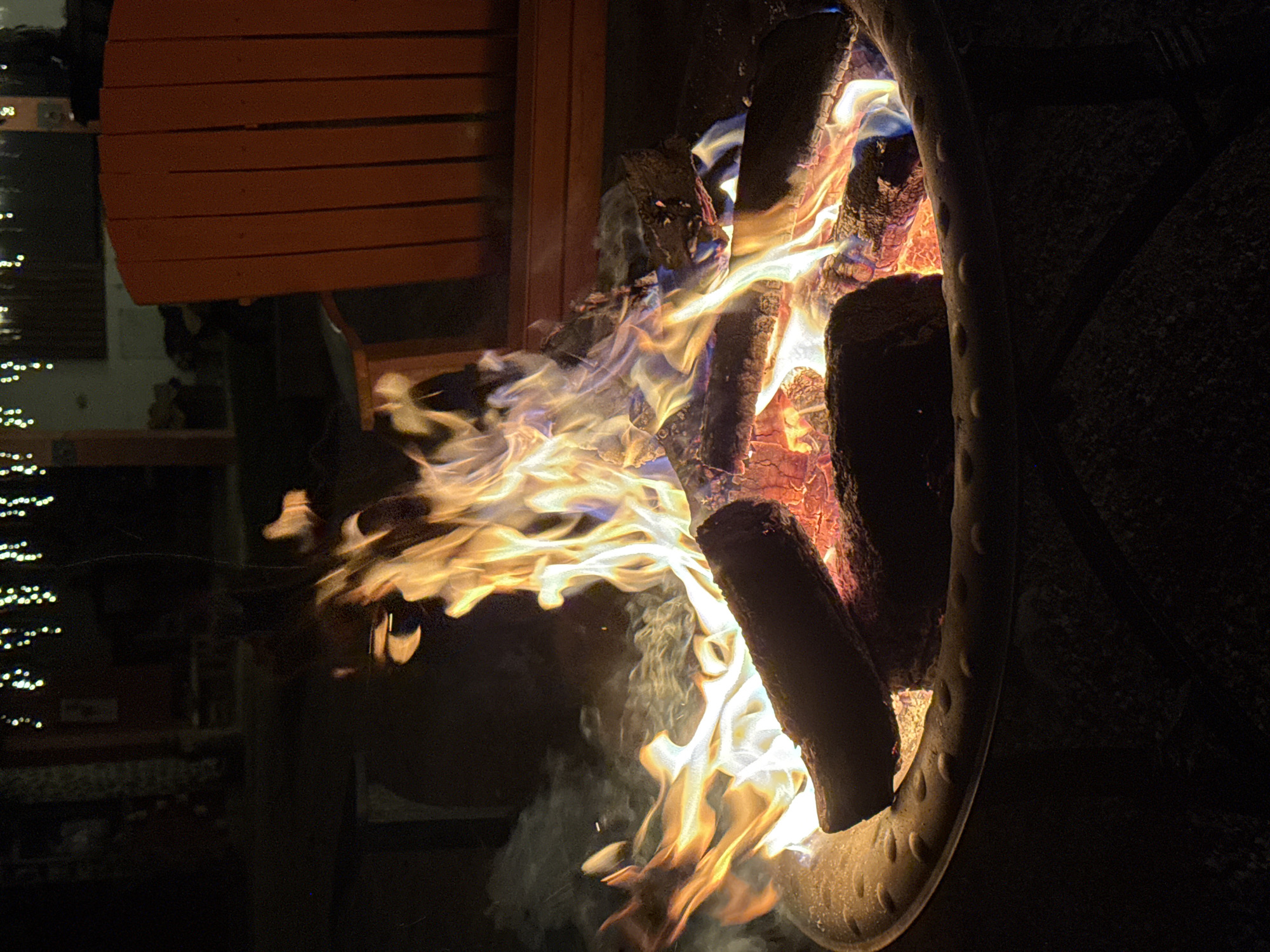 A close-up, low-light photograph shows a burning fire pit. Flames engulf logs within the metal fire pit ring, with smoke rising. To the left of the pit, part of an orange wooden chair is visible. In the blurred background, there are lights and a building. The location is presumed to be Cañada de Madero, Mexico, based on the prompt instructions. No people are visible.