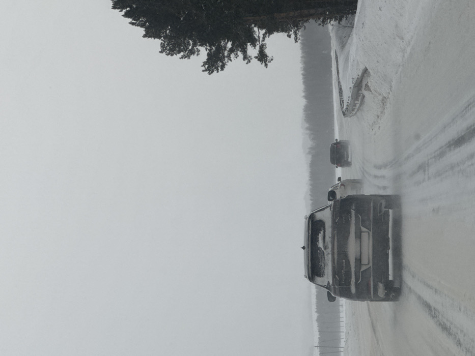 The scene depicts a winter road in Malyi Talmaz, Russia, under an overcast sky with snow covering the landscape. A dark-colored SUV, identified by faint markings as a Chery Tiggo 8 Pro Max, occupies the immediate foreground, its rear coated with snow on the window and tailgate, driving away from the viewer on a snow and slush-covered road with visible tire tracks. Following the SUV is a lighter-colored sedan, partially obscured. Further ahead, a dark-colored vehicle is in motion, generating a spray from its tires. The road curves slightly right, flanked by deep snowdrifts and a partially buried metal guardrail on the right side. A dense stand of coniferous trees lines the right edge of the road, while a distant treeline is visible across a snowy, open expanse in the background. No individuals are visible.