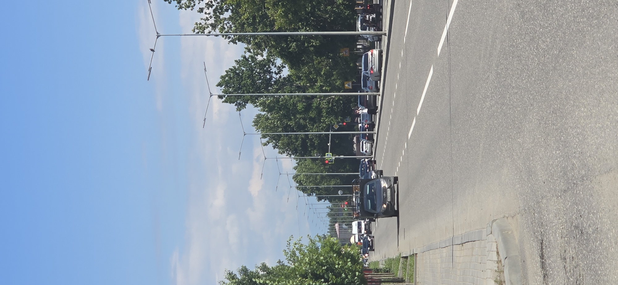 A vertical, eye-level shot captures a sunlit, wide asphalt road in Grozny, Russia, under a bright blue sky with scattered clouds. The road is lined with numerous tall streetlights on both sides, interspersed with lush green trees. Traffic is moderate, with several cars and a van visible. In the foreground, a dark-colored pickup truck with a black cap is moving forward, displaying a Russian license plate. To the left, a paved sidewalk runs alongside the road, bordered by a concrete curb. Further down the road, a traffic light is visible, indicating a controlled intersection. The scene appears to be an urban environment on a clear day.
