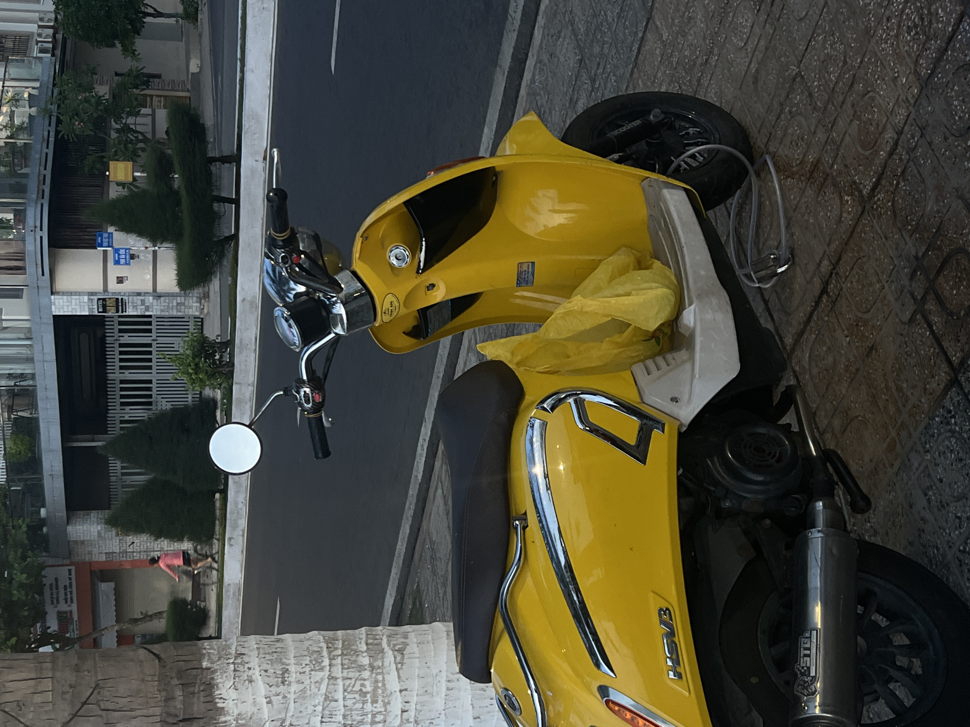 In the city of Bac Nha Trang Ward, Vietnam, a yellow scooter is parked on a sidewalk next to a street. The scooter has a black seat and a yellow plastic bag hanging from its handlebars. In the background, a person is riding a bicycle on the street, and buildings and trees line the street. The sidewalk is made of concrete tiles. The lighting suggests it is either early morning or late afternoon.