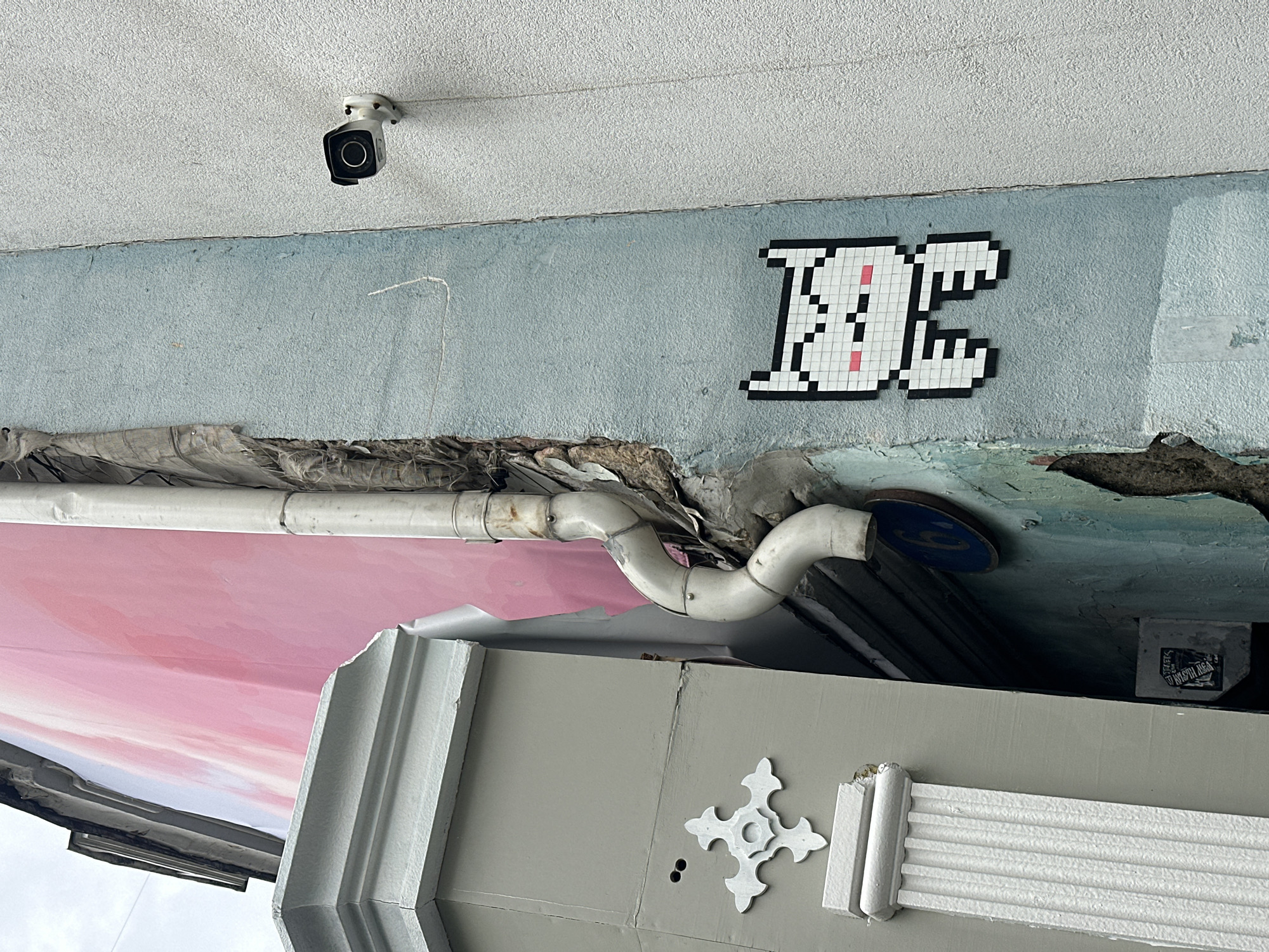 The image shows an outdoor scene in Unknown, Unknown, featuring a security camera mounted on a light gray ceiling. Below the ceiling, there's a light blue wall with a pixelated white and black "KC" design. A damaged section of the wall reveals exposed material, above which a white PVC pipe runs horizontally with bends. Below this, there is a fragment of a pink banner or awning. A gray structure with a decorative white cross and a fluted design is located at the bottom of the frame.