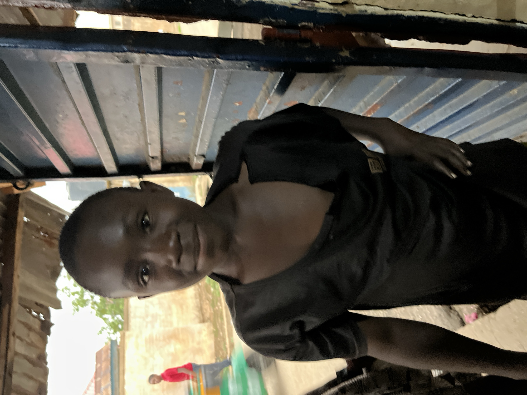 A person with dark skin looks up into the camera from under a blue metal structure. The person wears a dark shirt. In the background, a person wearing a red shirt stands near a green object. The location is possibly Jos, Nigeria, based on the context of the request. The environment appears to be urban with buildings and foliage visible.