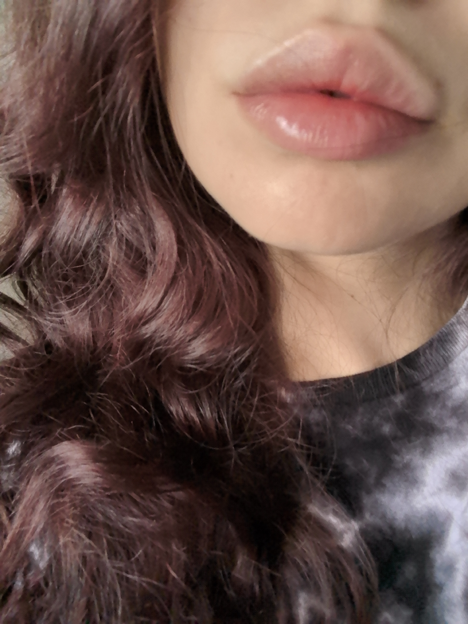 A close-up image depicts a portion of an individual's face and upper torso, situated indoors in the city of Stara Zagora, Bulgaria. The central focus is a set of full, naturally contoured lips exhibiting a pinkish hue and a noticeable sheen. The surrounding skin is fair-toned. To the left side of the frame and below the chin, dark reddish-brown hair with a distinct wavy or curly texture is visible. The individual is wearing a garment with a grey, black, and white tie-dye pattern, a portion of which is visible below the neck. No other individuals, specific objects, actions, or interactions are discernible within the frame.