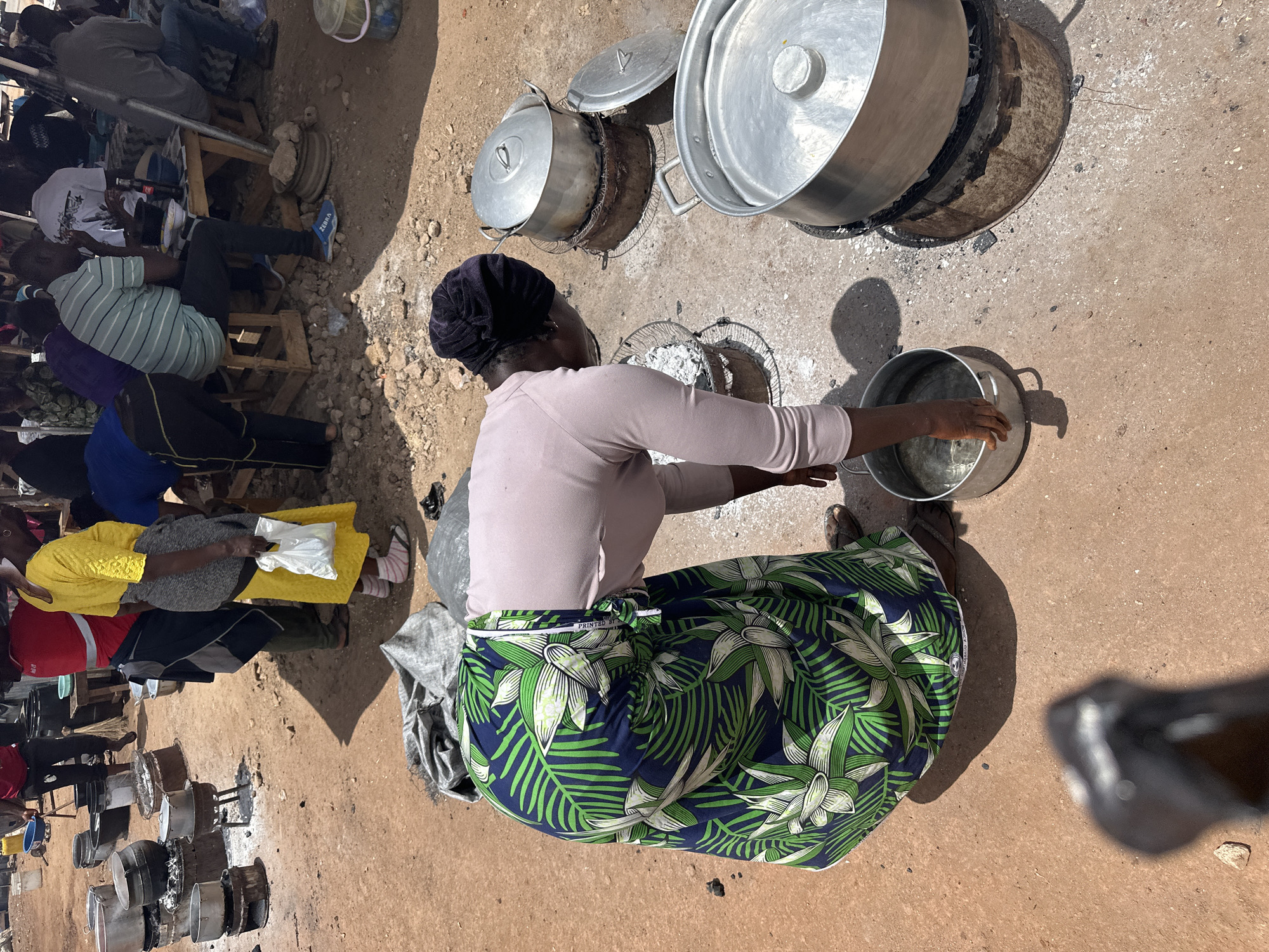 The image captures an overhead view of an outdoor cooking and gathering scene in Gura Top, Nigeria. The central subject is a woman, seen from above, wearing a light pink long-sleeved top and a vibrant blue and green patterned skirt (wrapper). She is actively engaged in handling a large silver pot, appearing to be stirring or adding contents to it.
Around her, the dusty, unpaved ground is dotted with multiple large aluminum cooking pots, many with lids, resting on what appear to be rudimentary charcoal stoves or open fires. Some pots show signs of being actively used for cooking, with ash visible around their bases. The sheer number of pots suggests large-scale food preparation, likely for communal consumption, a market, or an event.
In the background, several other individuals are visible, mostly seated on low wooden stools, observing or waiting. One person is wearing a striped shirt and blue sneakers. The scene is brightly lit by natural sunlight, indicated by the distinct, relatively short shadows cast by people and objects, suggesting it is midday or early afternoon. The weather appears dry and sunny.
Other objects include a white plastic bucket and a small plastic bag. On the patterned skirt of the main woman, the text "PRINTED BY" is visible. The overall atmosphere conveys a bustling outdoor environment focused on food preparation and social gathering.