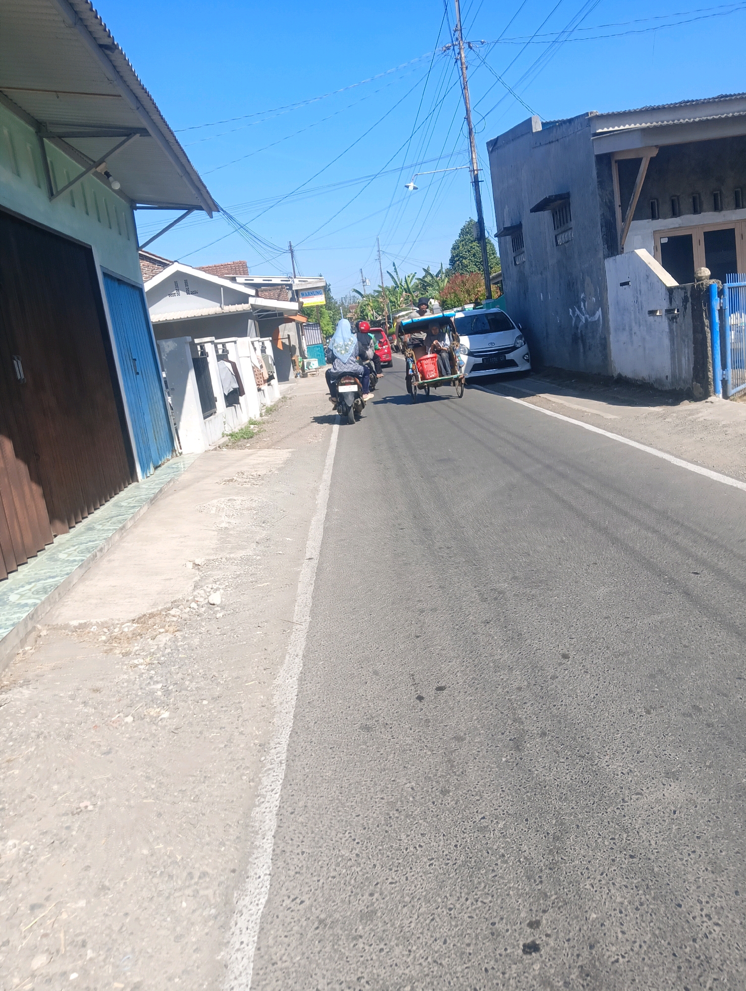 On a bright, sunny day in Sokaraja, Indonesia, a paved road runs through a residential area lined with houses and small businesses. A white car is parked to the right side of the road, partially obscured by a concrete wall. A motorcycle with two passengers, one wearing a blue headscarf, is seen in the middle of the road. Further ahead, a three-wheeled vehicle, also known as a "becak motor" or bemo, is driving with passengers. The buildings on the left side of the road appear to be shops or houses with corrugated metal roofs. Power lines crisscross the clear blue sky above.