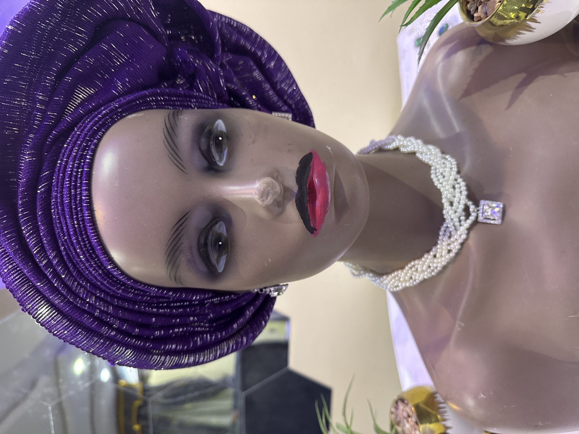A mannequin is adorned with a purple head wrap and a pearl necklace with a square, rhinestone clasp. The mannequin has painted features, including exaggerated eyebrows and painted lips. The background shows a hint of an interior setting. The location is Ajara, Nigeria.