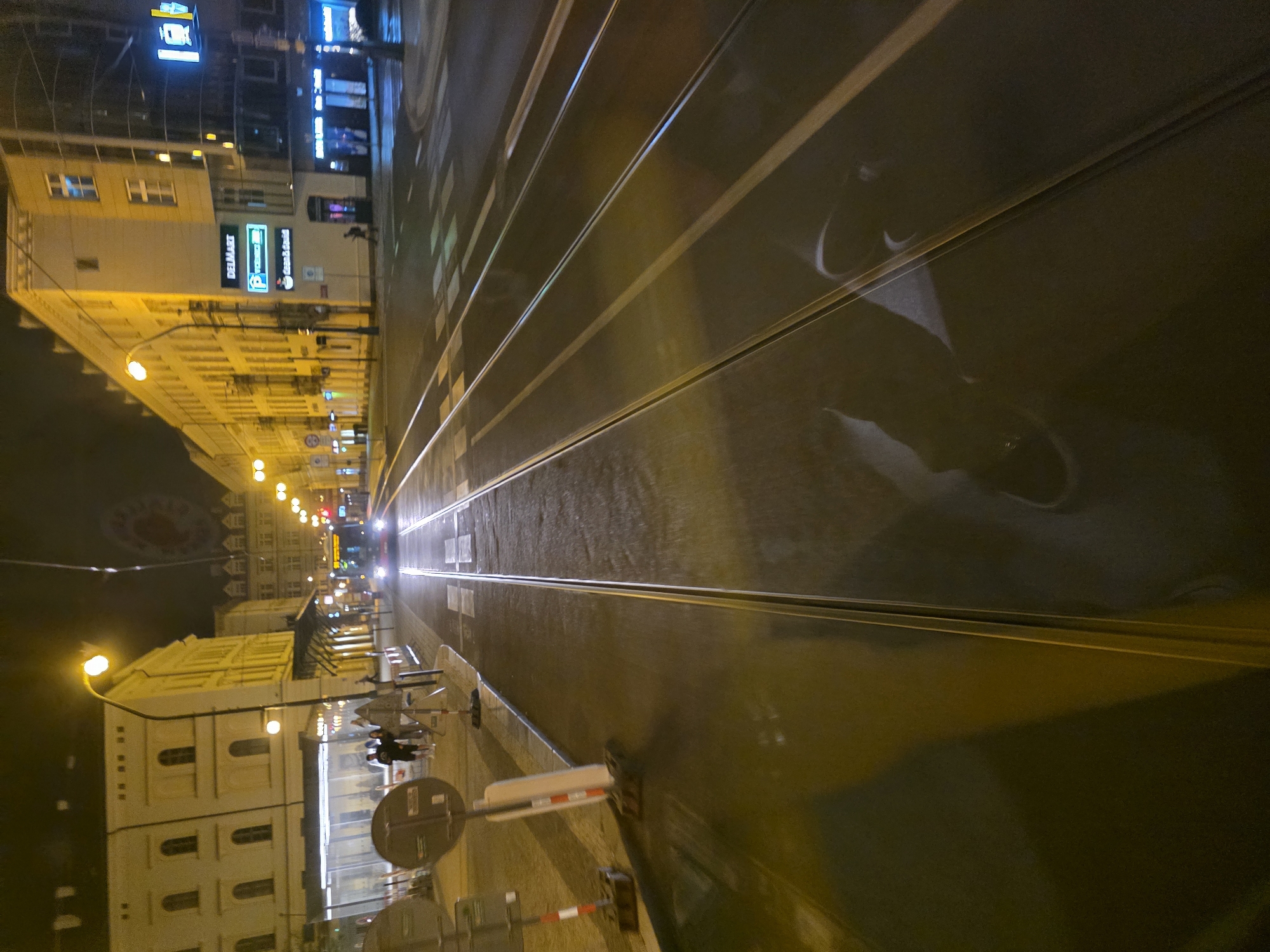 The image captures a tilted, low-angle perspective of a city street at night, looking down along tram tracks. The scene is illuminated by numerous streetlights and neon signs, casting a warm, yellowish glow on the buildings lining the left side of the street. The ground and tram tracks appear wet, with significant reflections of lights, buildings, and possibly a person's silhouette on the glossy surface, suggesting recent rain.
In the foreground and running diagonally through the center of the frame are prominent tram tracks. On the left, a row of multi-story buildings, primarily light-colored, stand under the artificial light. Several businesses are visible on the ground floors, identifiable by their illuminated signs. Notable among these is a light blue sign displaying "DEL MART" and another light blue sign with a white "P" for parking. Further down the street, a blue sign for "Česká spořitelna" (Czech Savings Bank) is visible, indicating the location is the Czech Republic.
Bright headlights of vehicles can be seen in the distance, moving along the street. Various street furniture, including traffic lights, road signs (one round with a red border and diagonal line, another plain round), and a barrier-like structure, are scattered along the pedestrian area and near the tracks. The overall atmosphere is that of a bustling urban environment late in the evening or at night.