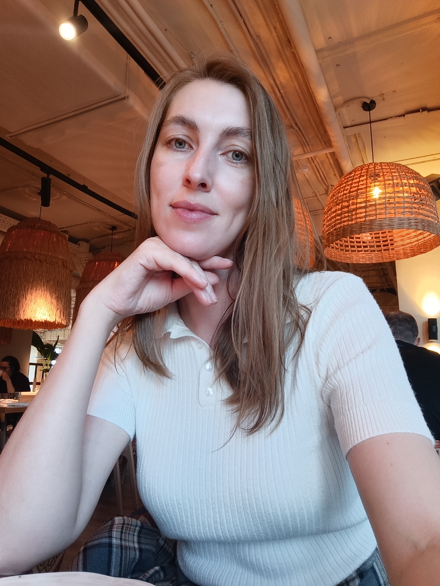 An adult female, appearing to be in her late 20s to early 40s, is seated indoors, looking directly at the camera with a subtle smile, her right hand supporting her chin. She has light skin, long light brown hair, and blue-green eyes. She is wearing a light-colored, ribbed knit polo shirt with a collar and three visible buttons, and a segment of a plaid garment is visible below her waist. The scene is an interior of a restaurant or cafe, specified as being in Moscow, Russia. The background features warm, amber lighting, exposed ceilings with white pipes, and suspended track lighting. Multiple large pendant light fixtures with woven rattan shades are visible, casting a soft glow. In the blurred background, two other individuals are partially visible: one person with dark hair seated at a table to the left, and another person seen from behind to the right. A green plant is also partially visible in the left background.