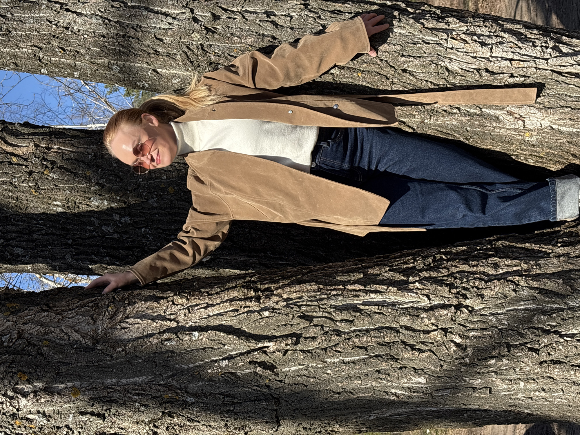 A blonde woman is depicted lying horizontally across a large, textured tree trunk in a natural outdoor setting, possibly a wooded area in Noviy, Russia. She is looking towards the camera with a gentle smile, her face illuminated by bright sunlight. She wears a light brown suede-like jacket, a white turtleneck shirt, dark blue jeans with cuffed ankles, and round, light-tinted sunglasses. Her hair is pulled back, revealing her profile. Her arms are extended, with her hands resting on the deeply furrowed bark at either side of her body.
The tree trunk beneath her is a prominent feature, showcasing intricate patterns of rough bark with varying shades of grey and hints of white or lichen. Above her, another substantial tree trunk creates a natural frame, through which a clear blue sky and sparse, bare tree branches are visible in the background. The bright sunlight casts distinct shadows on the tree bark, suggesting a sunny day, likely during mid-morning or afternoon. The scene conveys a relaxed and pleasant atmosphere. No text is visible in the image.