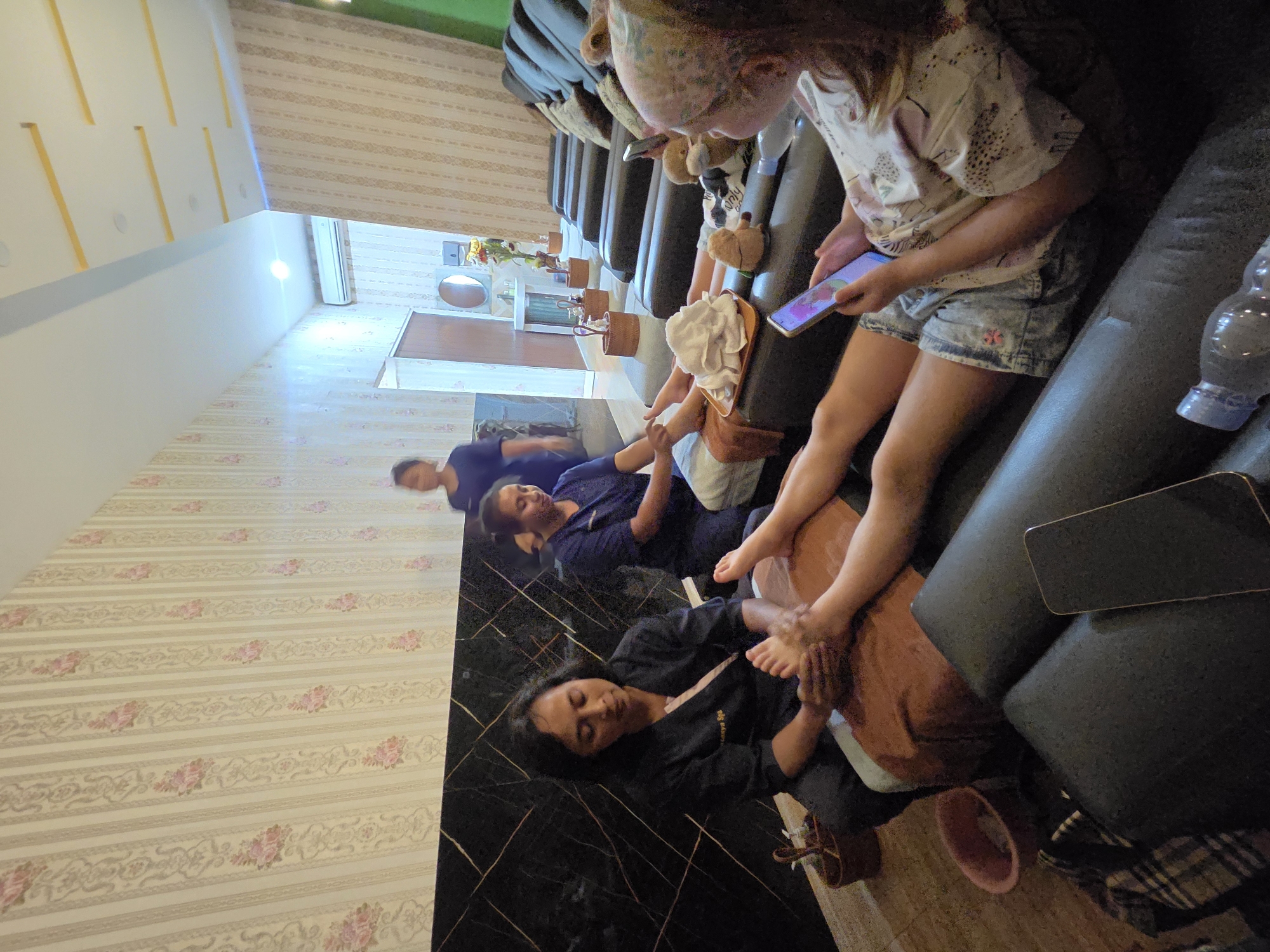 The image shows an indoor spa or massage parlor in North Kuta, Indonesia. Three women are visible. In the foreground, a woman in a dark uniform is massaging the foot of another woman who is lying down on a dark-colored couch, holding a mobile phone. The woman receiving the massage is wearing denim shorts and a patterned t-shirt. Further back, another woman in a similar uniform is attending to another client, whose legs are visible, also lying on a couch. In the background, a third woman in a dark blue top stands and appears to be observing or waiting. The room features a wall with floral wallpaper and dark, patterned accents. There are multiple reclining couches or massage chairs arranged in a row, suggesting a shared treatment space. Baskets containing towels and other spa items are placed around the room. A water bottle is visible in the lower right corner.
