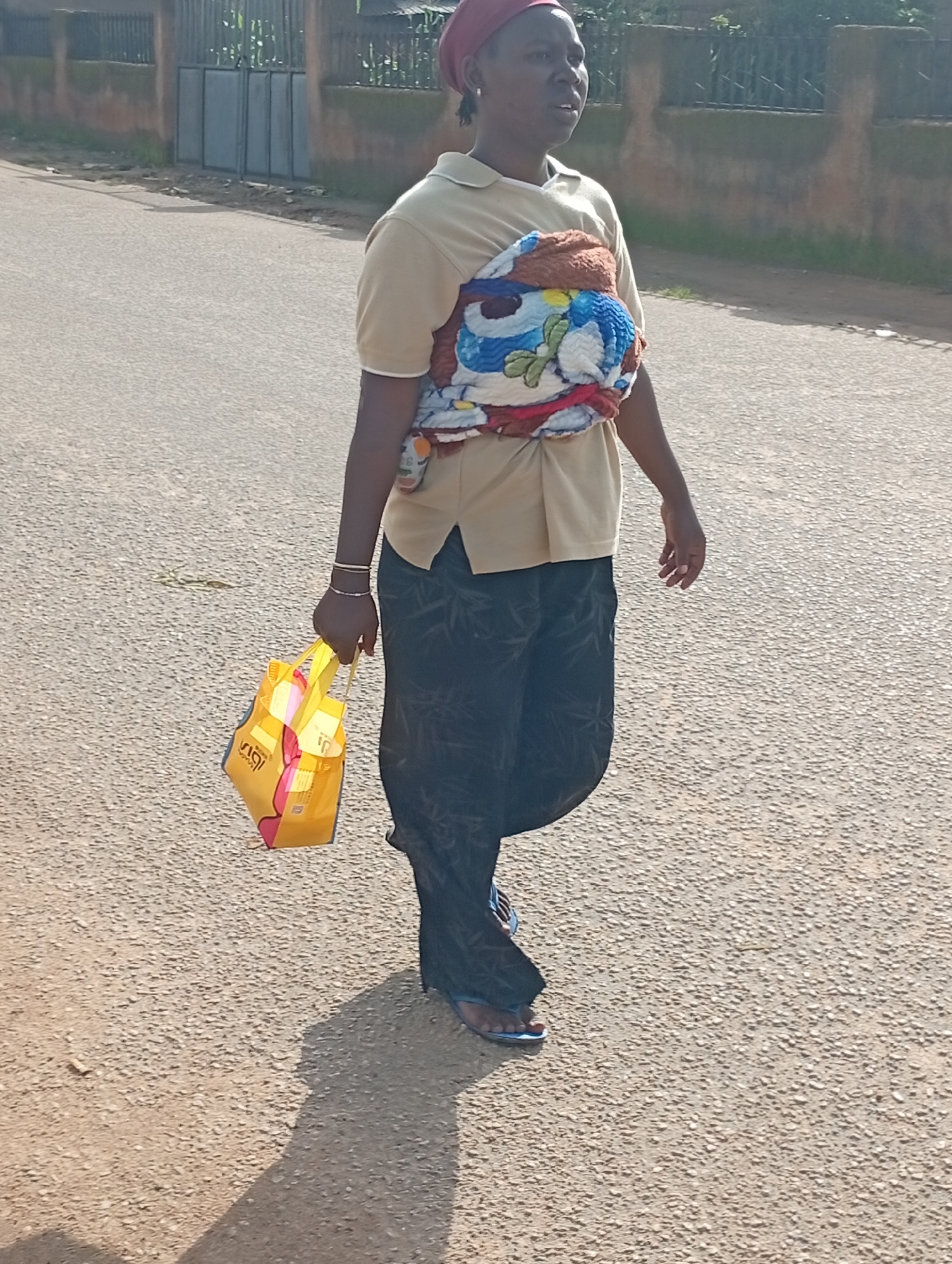 A person is walking on a paved road in Jos, Nigeria. The person is carrying a baby wrapped in a colorful blanket on their back and holding a yellow shopping bag. The person is wearing a light-colored short-sleeved shirt, dark patterned pants, and blue flip-flops. In the background, there is a fence and some vegetation. The sun is casting a shadow on the road.