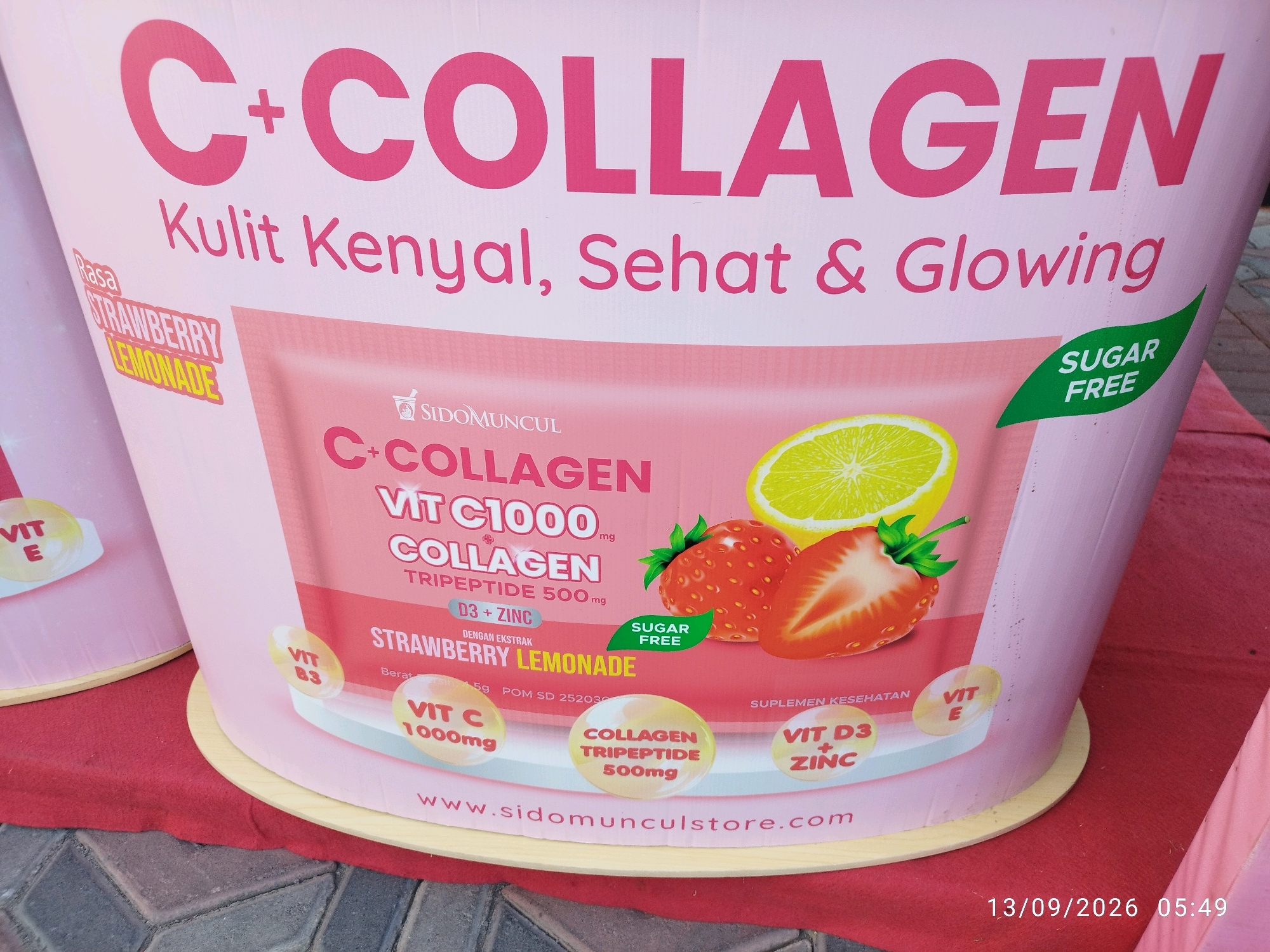 An outdoor advertising display featuring a large, pink cylindrical object with the prominent text "C+COLLAGEN" in bold pink lettering. Below this, smaller text reads "Kulit Kenyal, Sehat & Glowing" (Supple, Healthy & Glowing Skin). To the left, a pink label with "Rasa STRAWBERRY LEMONADE" is visible. The main display showcases a stylized pouch of "C+COLLAGEN VIT C1000 COLLAGEN TRIPEPTIDE 500mg" with an image of strawberries and a lemon slice. A green leaf shape with "SUGAR FREE" is also present. Various circular icons highlight "VIT E", "VIT B3", "VIT C 1000mg", "COLLAGEN TRIPEPTIDE 500mg", and "VIT D3 + ZINC". The bottom of the display features the website "www.sidomunculstore.com". The object is situated on a red cloth covering a surface that appears to be a cobbled walkway or pavement. The timestamp "13/09/2026 05:49" is present in the lower right corner, indicating the date and time the photograph was taken. The scene is likely in a public area or market, possibly within the city of Mojokerto, Indonesia, given the context of local Indonesian products. No individuals are visible in the image.