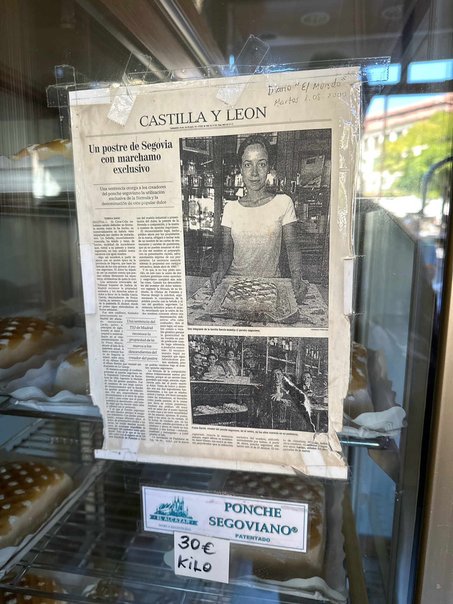 A close-up, eye-level, outdoor shot shows a newspaper clipping taped to the inside of a display window. The newspaper clipping, dated Tuesday, August 1, 2000, features a headline in Spanish reading "A dessert from Segovia with exclusive marchamo." The article discusses a court ruling granting exclusive rights to the creators of "ponche segoviano," a popular dessert. A black and white photograph shows a woman presenting a ring-shaped dessert. Below this, another photograph depicts several people gathered around a table, with one individual holding a similar dessert. At the bottom of the clipping, a label for "EL ALCAZAR" and "PONCHE SEGOVIANO PATENTEADO" is visible, along with pricing information of "30€ KILO." The display window is likely in La Lastrilla, Spain, given the newspaper's focus on a Segovian dessert.