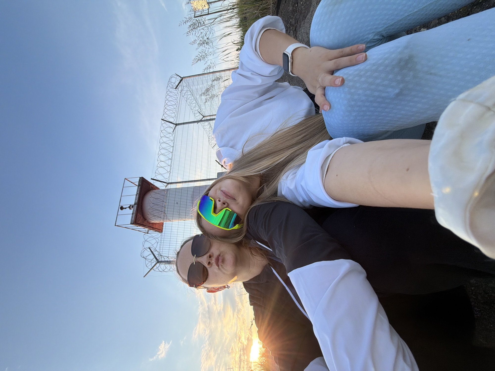Two women are posing for a selfie outdoors. The woman in the foreground on the left is wearing large sunglasses and pursing her lips. She is wearing a black hoodie with white sleeves. The woman on the right is leaning her head on the other woman's shoulder and wearing bright, colorful goggles. She is wearing a white shirt and light blue patterned pants. A lighthouse and a fence with barbed wire are visible in the background, along with tall grass. The sun is setting, casting a warm glow on the scene. The location is near the Kama River in Chaikovsky, Russia.