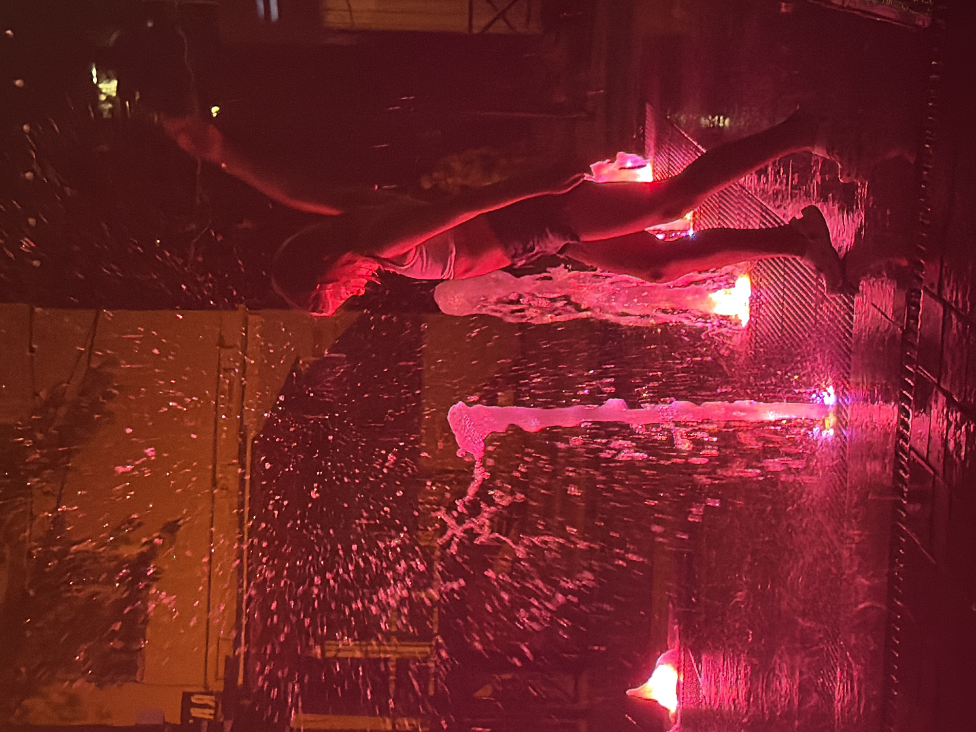 The image depicts a person, likely an adult, in the process of performing a water-based act, possibly a dive or a jump, into a body of water. They are captured mid-air, with their body extended horizontally and their legs slightly bent. The person is wearing what appears to be a swimsuit or shorts.
The scene is illuminated by bright pinkish-red lights, creating a dramatic and atmospheric effect. Water is being sprayed or is splashing around the person, indicating an active water feature or performance. The water itself seems to be illuminated by the red light, creating glowing trails and droplets.
The setting appears to be an outdoor area, possibly a pool or a water park, with a tiled or textured surface visible beneath the water. In the background, there are hints of structures and possibly some foliage, but they are obscured by the lighting and water effects. The time of day is difficult to determine precisely due to the artificial lighting, but the overall mood suggests an evening or nighttime event.
The activity suggests a performance or an intentional display of skill involving water. The bright, saturated lighting and the dynamic action of the water create a sense of excitement and visual spectacle. There are no visible text elements in the image. The location context of Partenit, Ukraine is provided, but not directly evident from visual cues within the image itself.