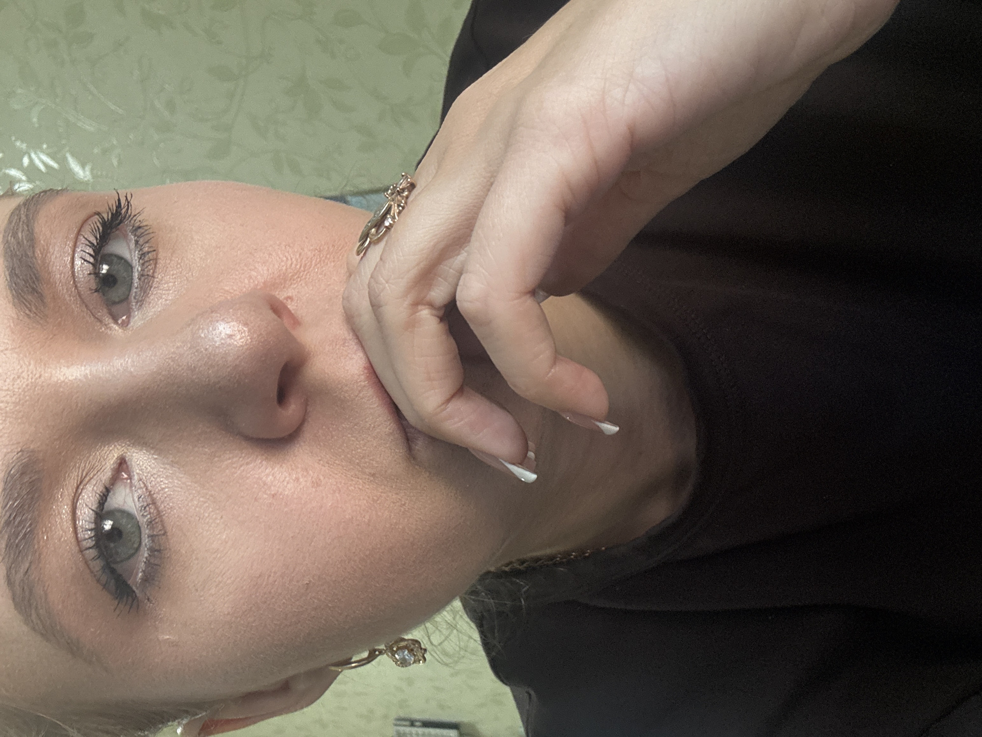 A close-up portrait of a young Caucasian woman's face and upper body is shown. She is looking towards the viewer with her right eye and nose in focus. Her left hand is touching her chin, and she is wearing a gold ring with a butterfly design on her right index finger. She has green eyes and her eyebrows are filled in. She is wearing a dark brown shirt and a gold earring with a white stone in her left ear. The background is a green floral wallpaper. The image appears to be taken indoors.