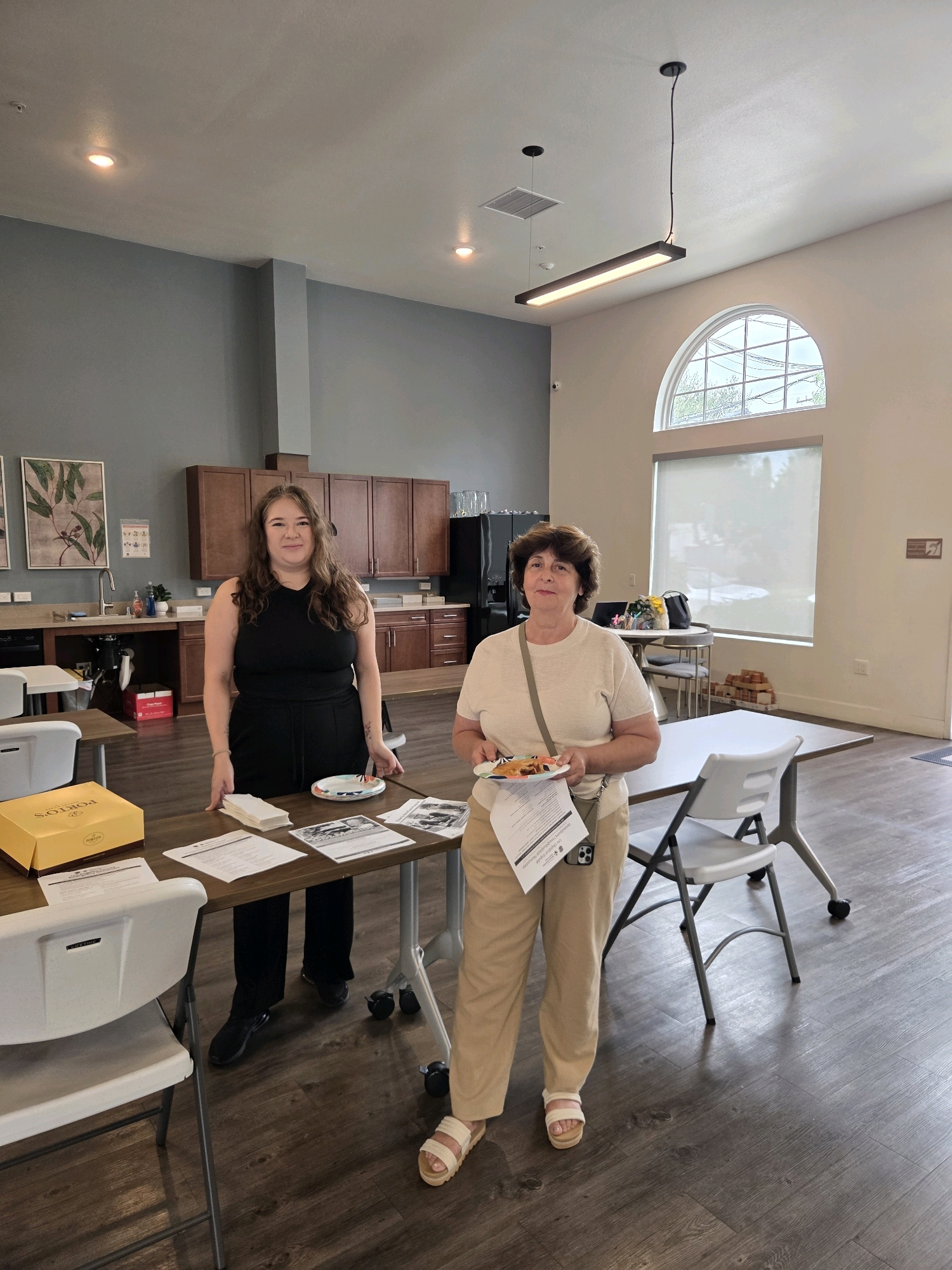 Two women stand in a common area that appears to be part of an apartment complex or community center in Glendale, United States of America. The room has a kitchenette area with wooden cabinets and a sink on the left side of the image. The floor is made of dark wood-look laminate.
The woman on the left is wearing a black sleeveless top and black pants. She has long brown hair and is looking at the camera with a slight smile. She is standing behind a wooden table with several papers and a yellow box on it. A small plate with what appears to be food is also on the table in front of her.
The woman on the right is wearing a beige short-sleeved shirt and tan pants. She has short brown hair and is holding a plate with food and a paper in her left hand. She is also looking at the camera. She is standing in front of a white table with a chair.
In the background, there is a large arched window with blinds. A long, modern light fixture hangs from the ceiling. The walls are painted a light color, with one wall on the left being a darker gray. Artwork depicting plants is visible on the gray wall. Several chairs and tables are arranged in the room.