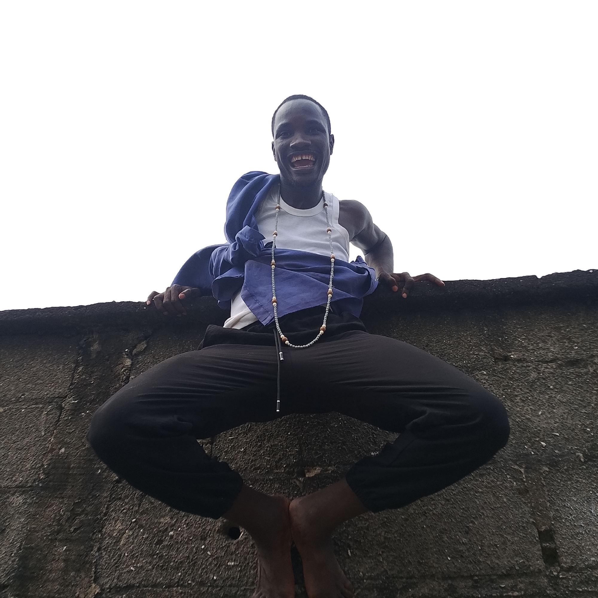 A low-angle, full-shot photograph captures a Black male sitting on a concrete wall with his legs spread wide. He is wearing a white tank top, a blue shirt draped over his shoulders, and black pants. A long, beaded necklace hangs from his neck. His head is tilted back, and he is laughing with his mouth wide open. His arms are resting on the wall, with his hands visible. The background is a bright, overcast sky. The concrete wall appears textured and aged, suggesting it might be outdoors in a somewhat weathered urban environment. This scene could be located in Ado, Nigeria, given the description.