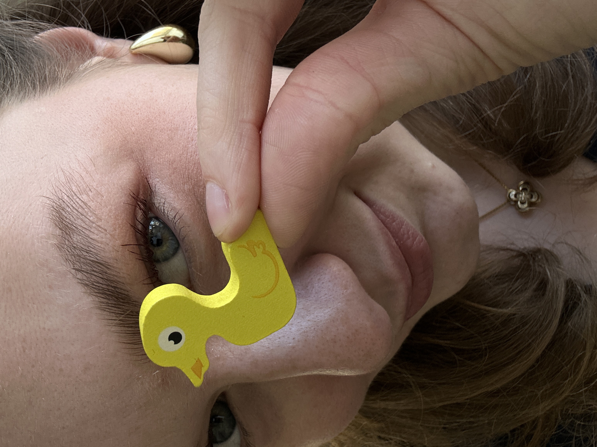 A close-up, slightly rotated, image features a fair-skinned person with light brown hair. The person is holding a small, yellow, wooden duck toy near their face. The toy's eye and beak are visible. The person's right eye is visible, and they have long eyelashes. A gold earring is visible on the person's right ear, and a gold necklace with a clover-shaped pendant is visible near their neck. There is no indication of the location being in Kazan, Russia.