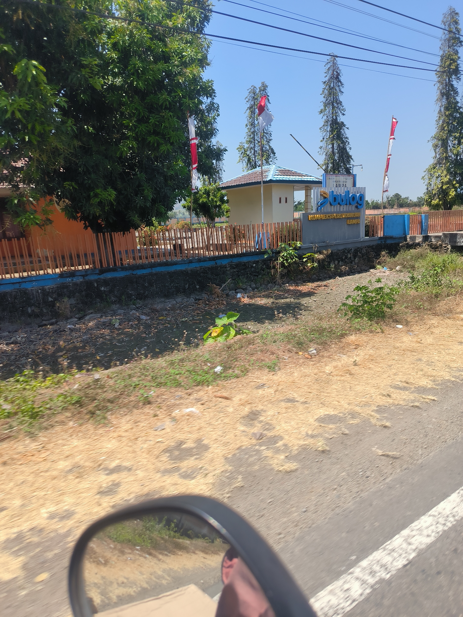 This is a medium shot, eye-level, outdoor, daytime, photograph of a government building in Pinrang, Indonesia. The building has a tan facade with a light blue roof. Above the building, a sign reads "BULOG" in large blue letters, with smaller text beneath it. Two Indonesian flags are flying from poles in front of the building. A wooden fence runs along the perimeter of the property. The surrounding environment is dry and arid, with sparse vegetation and cracked earth. In the foreground, a motorcycle's rearview mirror reflects a portion of the scene.