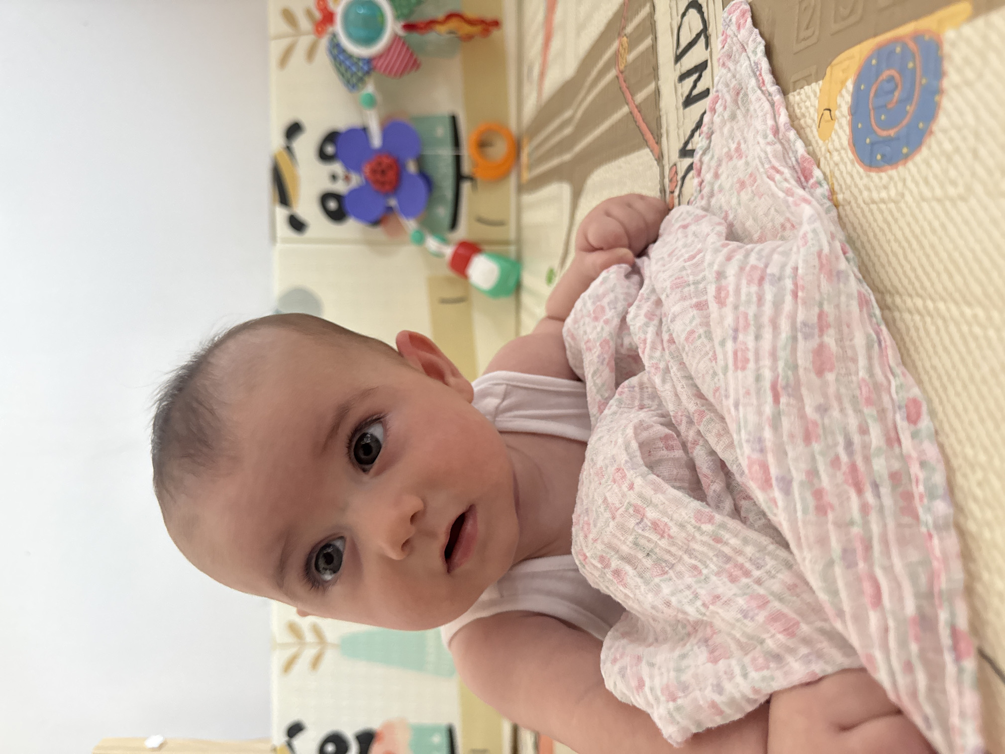 The image shows a close-up of a baby lying on a playmat. The baby is wearing a white tank top and is partially covered by a light pink blanket with a floral pattern. The baby's head is turned to the side, looking upwards and to the left, with a slightly open mouth. In the background, there are colorful baby toys attached to a play gym, including a panda-themed toy with purple and blue elements. The playmat has a patterned design that includes what appear to be roads and some circular shapes with spiral designs. The image quality is good, with clear focus on the baby and the immediate surroundings. The scene is indoors, likely a nursery or play area. The location of Isola delle Femmine, Italy, is not discernible from the visual information provided in the image.