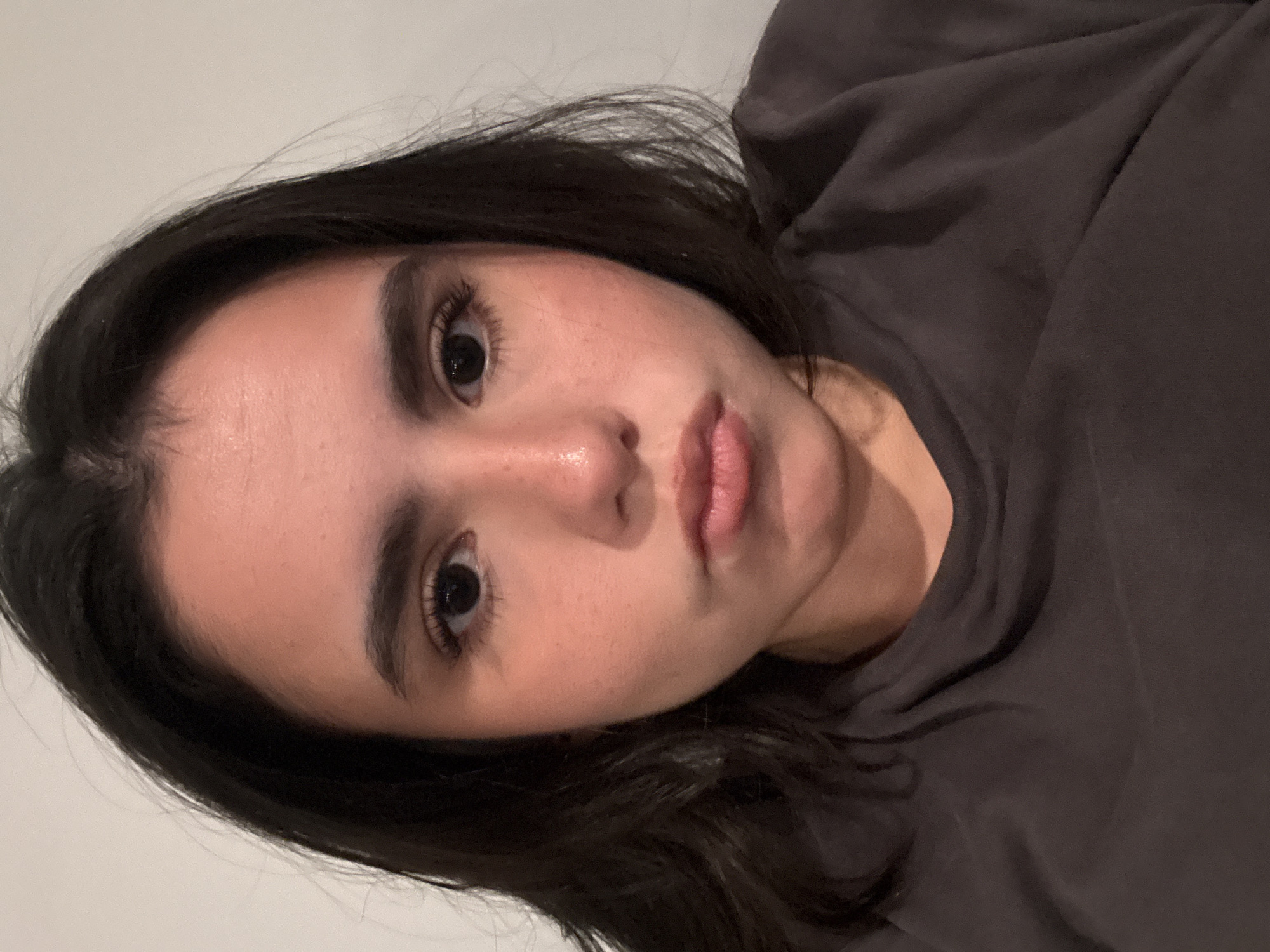 A young woman with dark hair is seen in a close-up, slightly rotated view. Her skin tone appears fair with some visible freckles. She has dark eyebrows and eyelashes, and her eyes are dark brown. Her lips are pink and full. She is wearing a dark gray or brown top that is slightly draped. The background appears to be a plain, light-colored wall. The location is not discernible from the image and cannot be determined to be in Austin, United States of America.