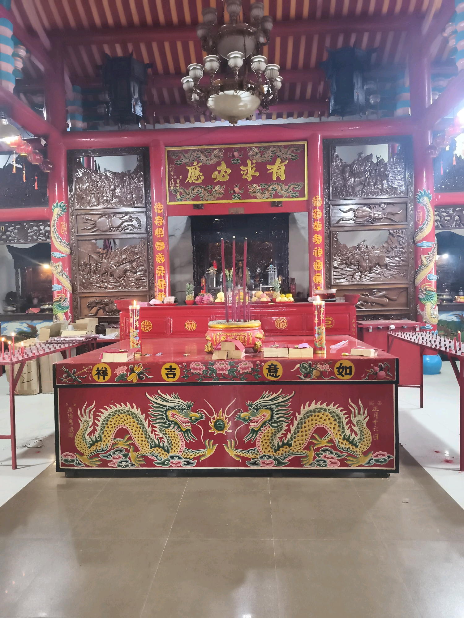 The image depicts the interior of a Chinese temple, likely located in Palembang, Indonesia, characterized by its vibrant red and gold color scheme. Dominating the foreground is a large, red altar adorned with intricate carvings of dragons. The altar is set with numerous lit incense sticks in a golden censer, offerings of fruit, and stacks of paper. On a lower level of the altar, there are more offerings and a single lit candle.
Behind the main altar, another red structure serves as a backdrop, featuring a framed inscription with Chinese characters and ornate wooden carvings on either side. These carvings appear to depict scenes with figures. Flanking this central display are tall, red pillars with further carvings, including a dragon winding its way up the left pillar. The ceiling is a striking striped pattern of red and yellow, with a large, ornate chandelier hanging centrally. The floor is tiled in a neutral, reflective material. Red lanterns and other decorative elements are visible throughout the space, contributing to the festive and spiritual atmosphere. The overall impression is one of a well-maintained and active place of worship.
